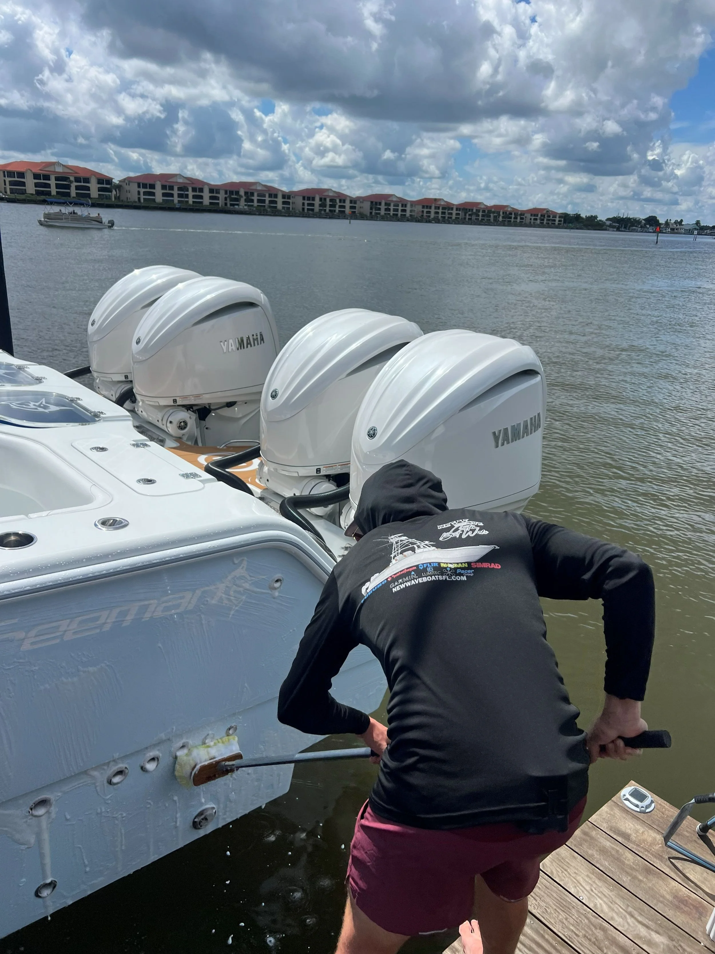 Person cleaning the bottom of a boat at a marina, with four Yamaha outboard motors in the background and water and buildings in the distance.