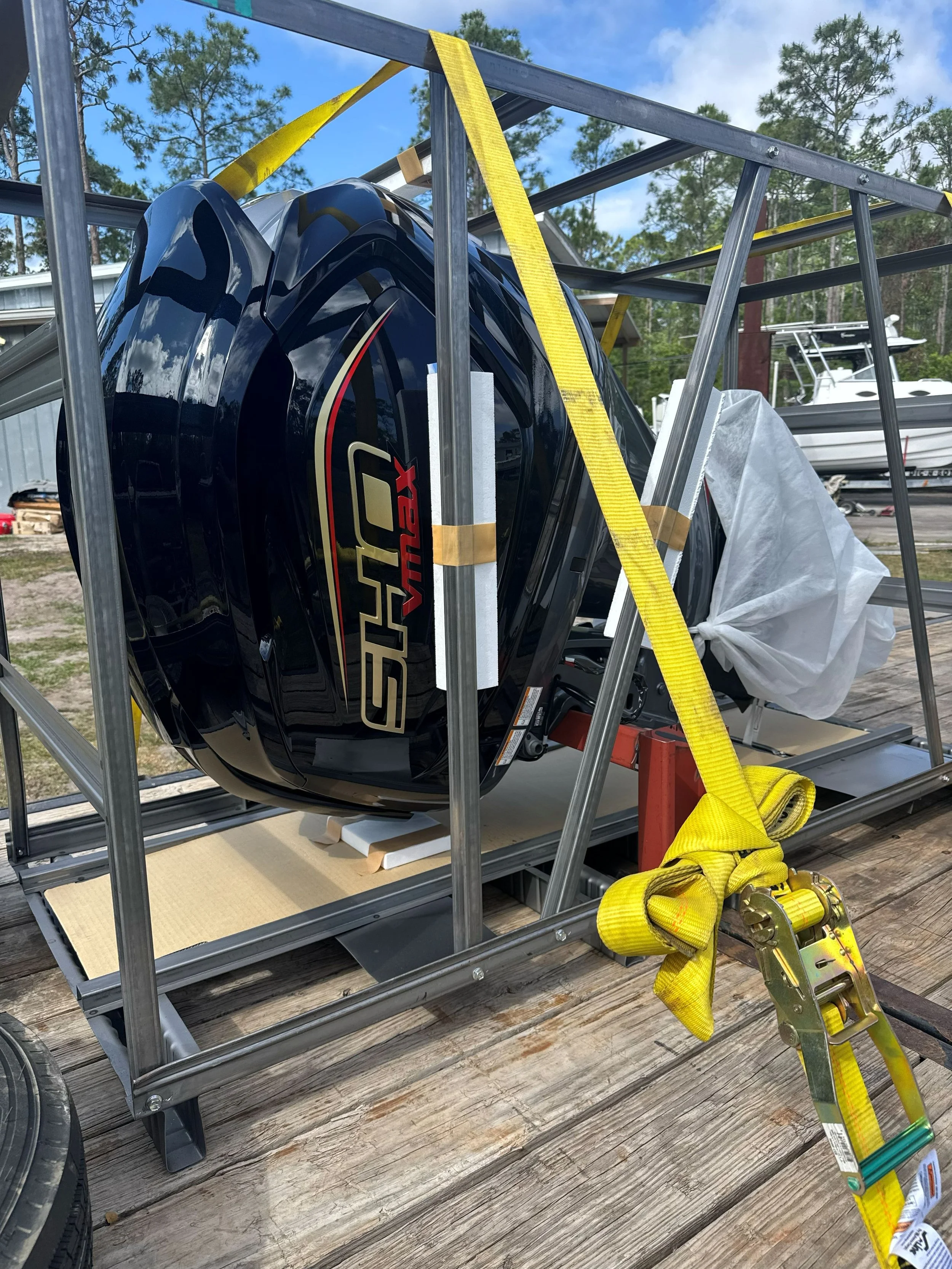 Outboard motor on a metal stand with yellow securing straps, fishing boats in the background, outdoors on a wooden surface.