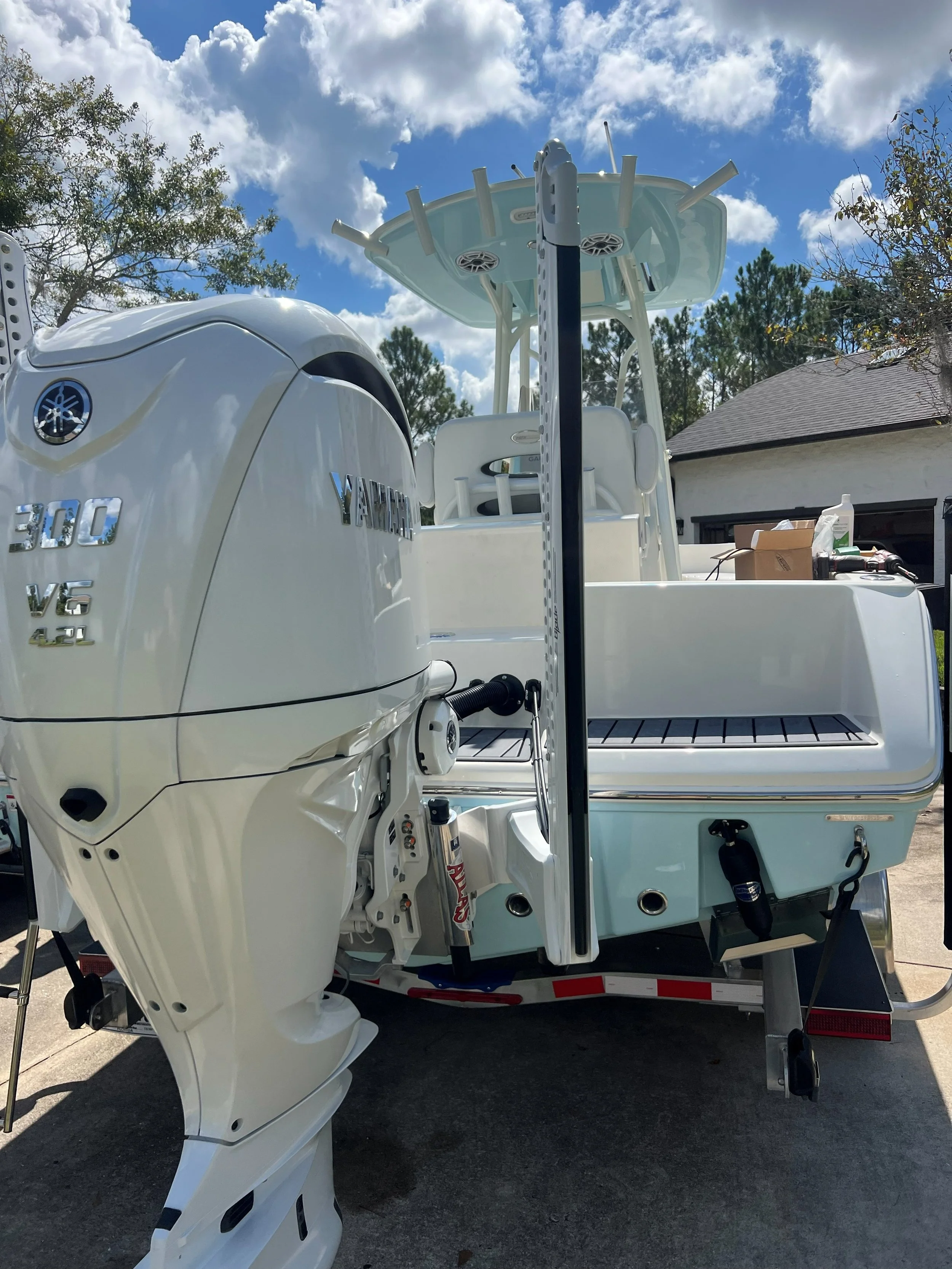 A white Yamaha outboard motor attached to a boat with a boat console, seating area, and T-top, parked on a trailer in driveway under partly cloudy sky.