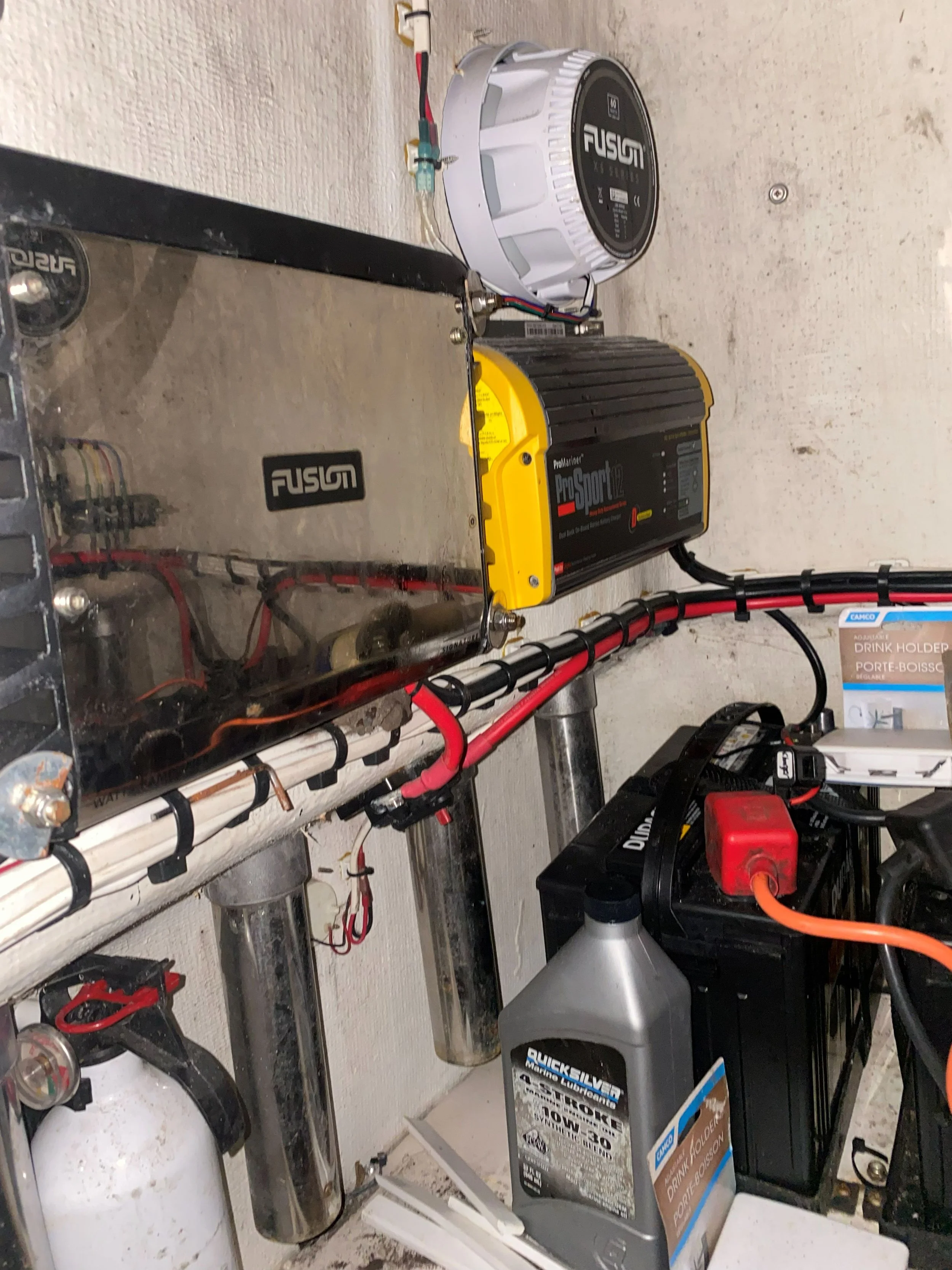 Electrical components and equipment mounted on a wall, including a Fusion power generator, a yellow PowerSport ProSport 12 inverter, a white Fusion speaker, a black battery, a red and black power cord, and various wires and tools.