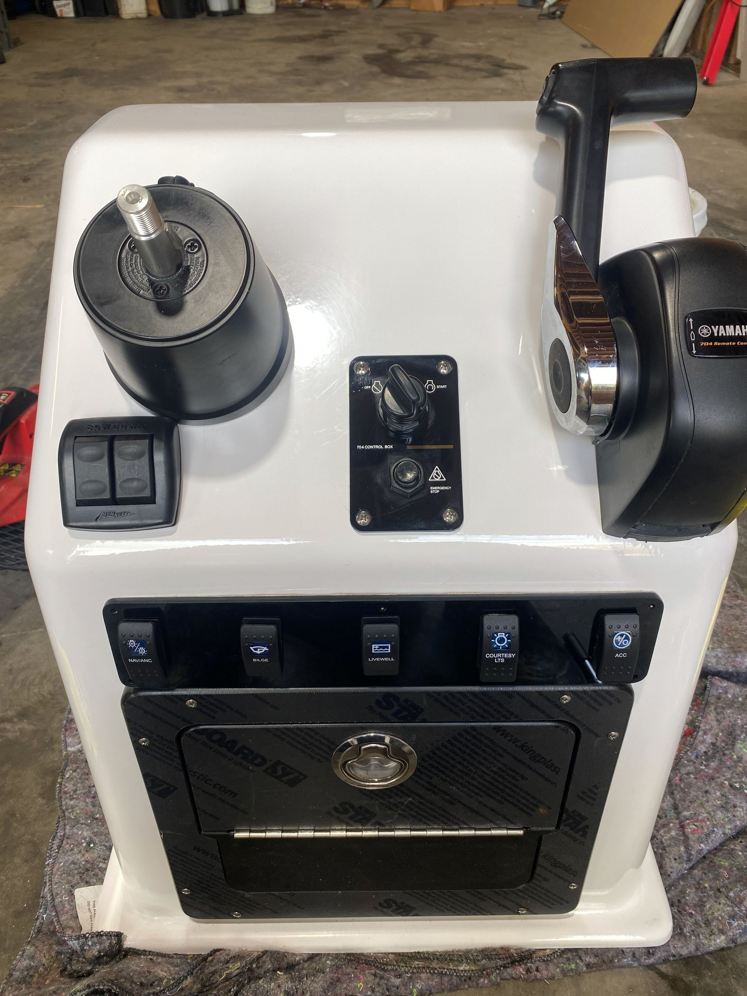A white marine vessel control console featuring a black steering wheel with a chrome hub, a black throttle handle, and various control switches and gauges on a black panel.