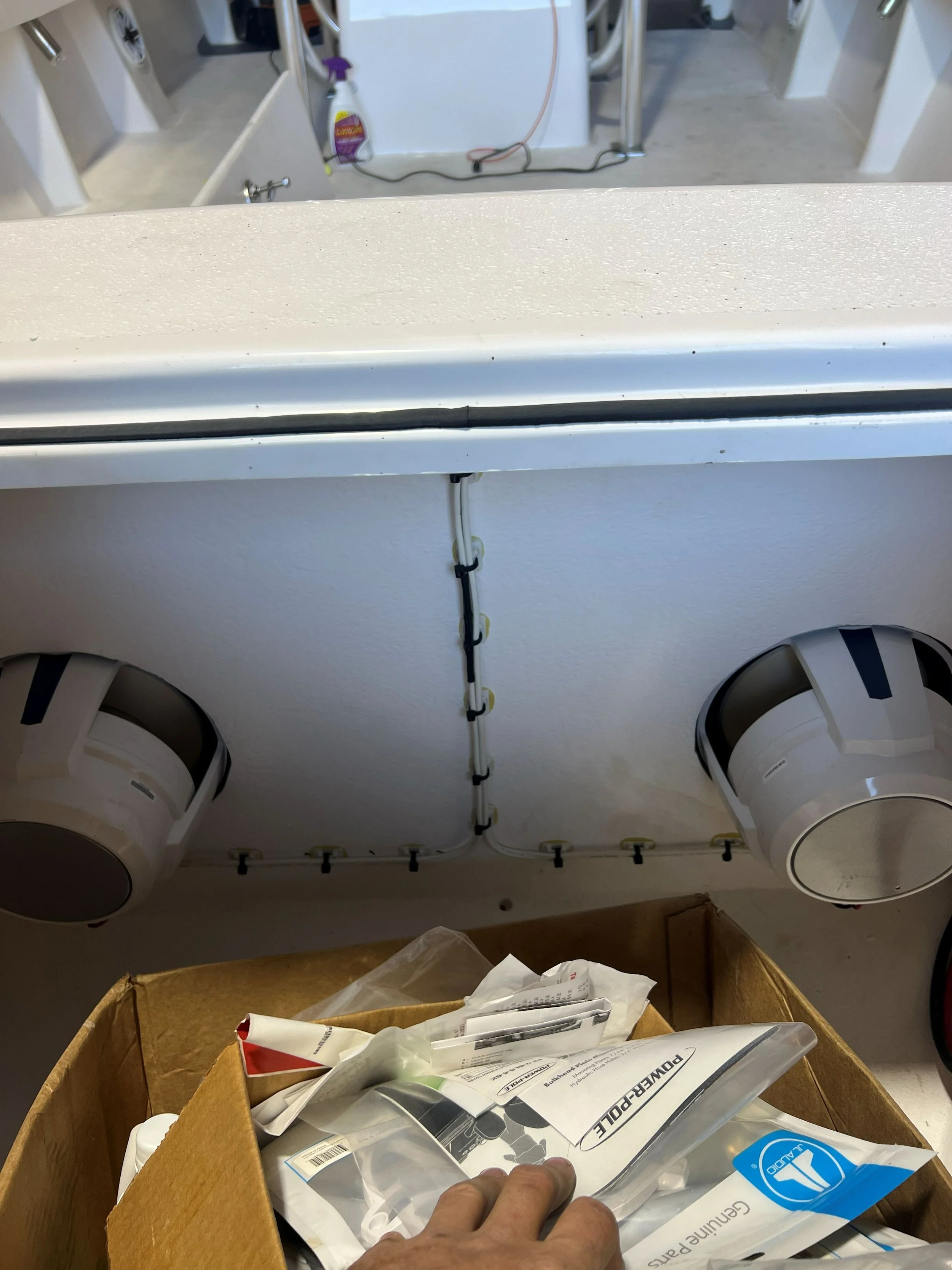 View of a laundry or utility room with a countertop, exposed plumbing, and two large round vents or exhaust fans on the ceiling. There is a cardboard box filled with various items and a person's hand in the foreground.