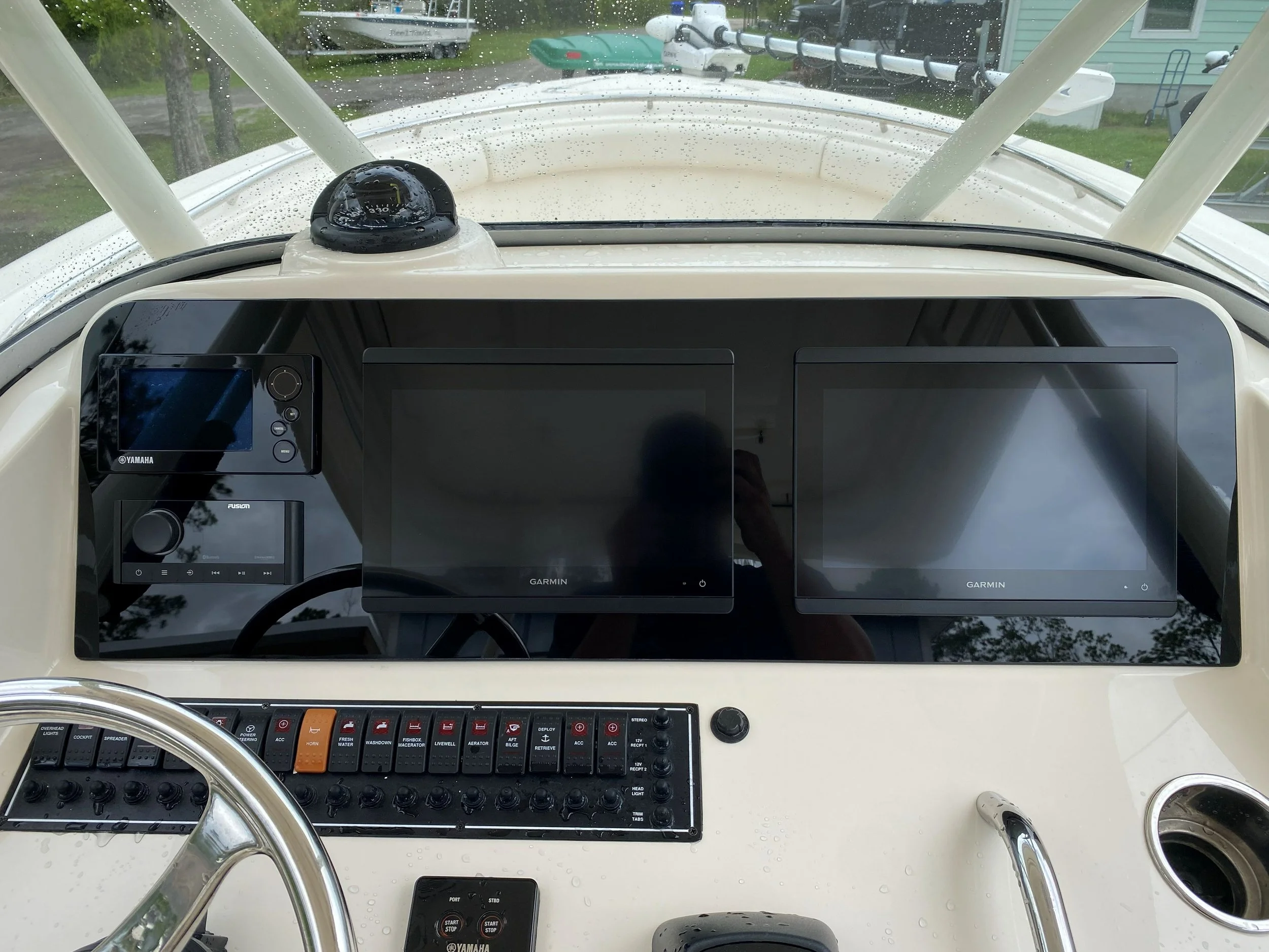 Dashboard of a boat with three large Garmin screens, control switches, a steering wheel, and a compass on top, with rain droplets on the windshield.
