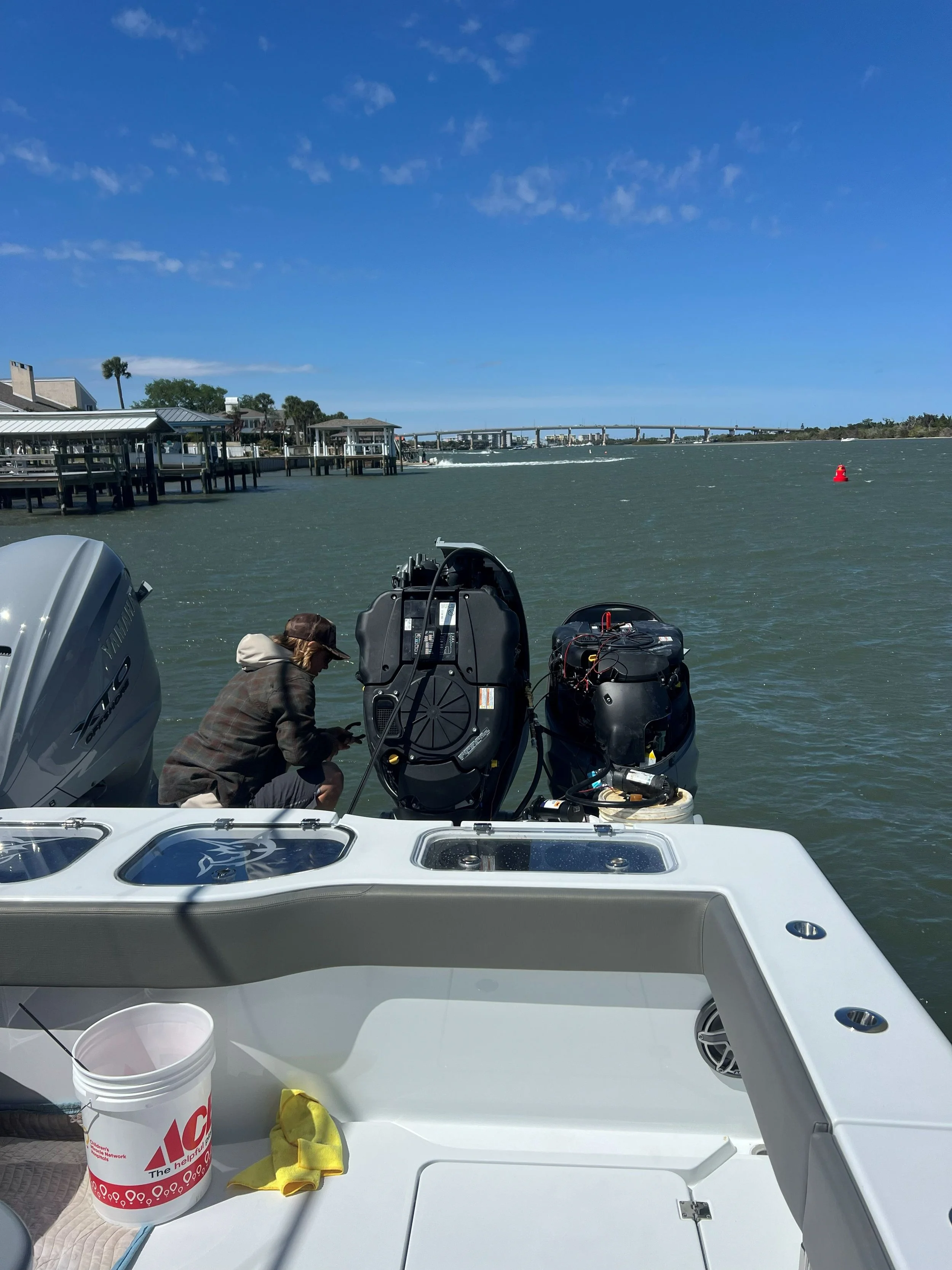 A person on a boat working near the water with outboard motors, with a shoreline, docks, and a bridge in the background.