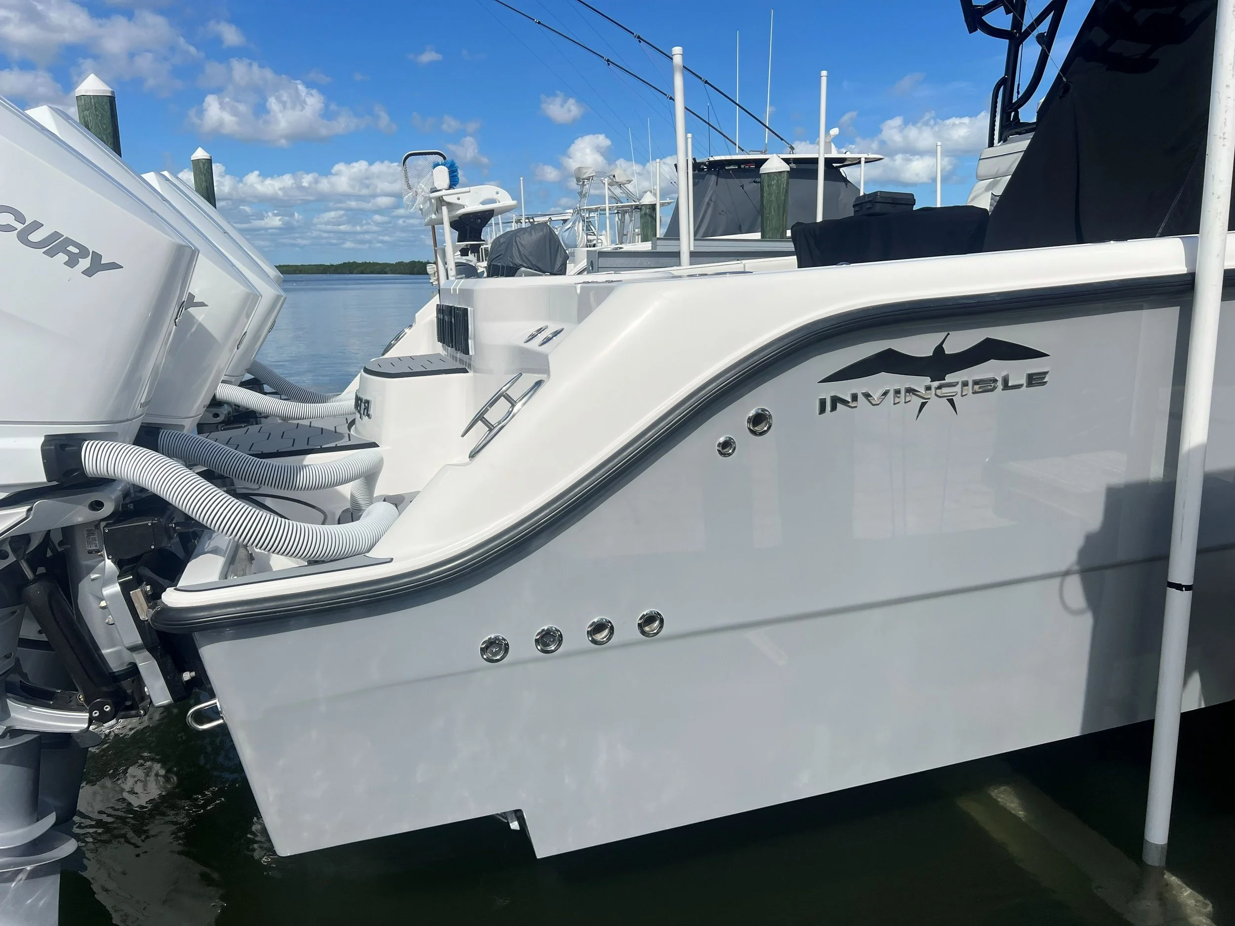 A white boat docked at a marina with the name Invincible on its side, and a Mercury engine attached at the back, under a partly cloudy sky.