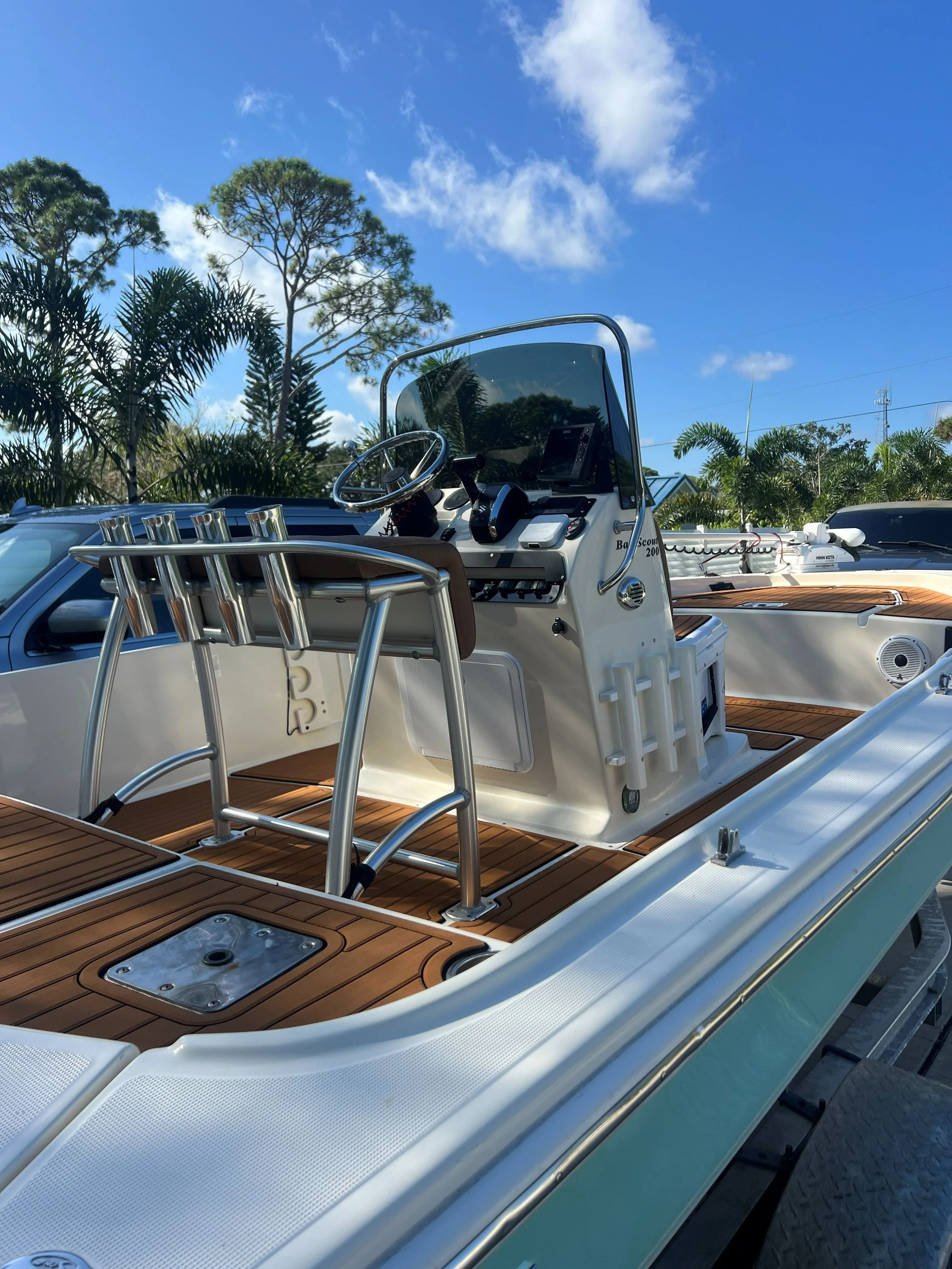A white boat with a wooden deck, metal railing, and steering console, parked outdoors with trees and blue sky in the background.