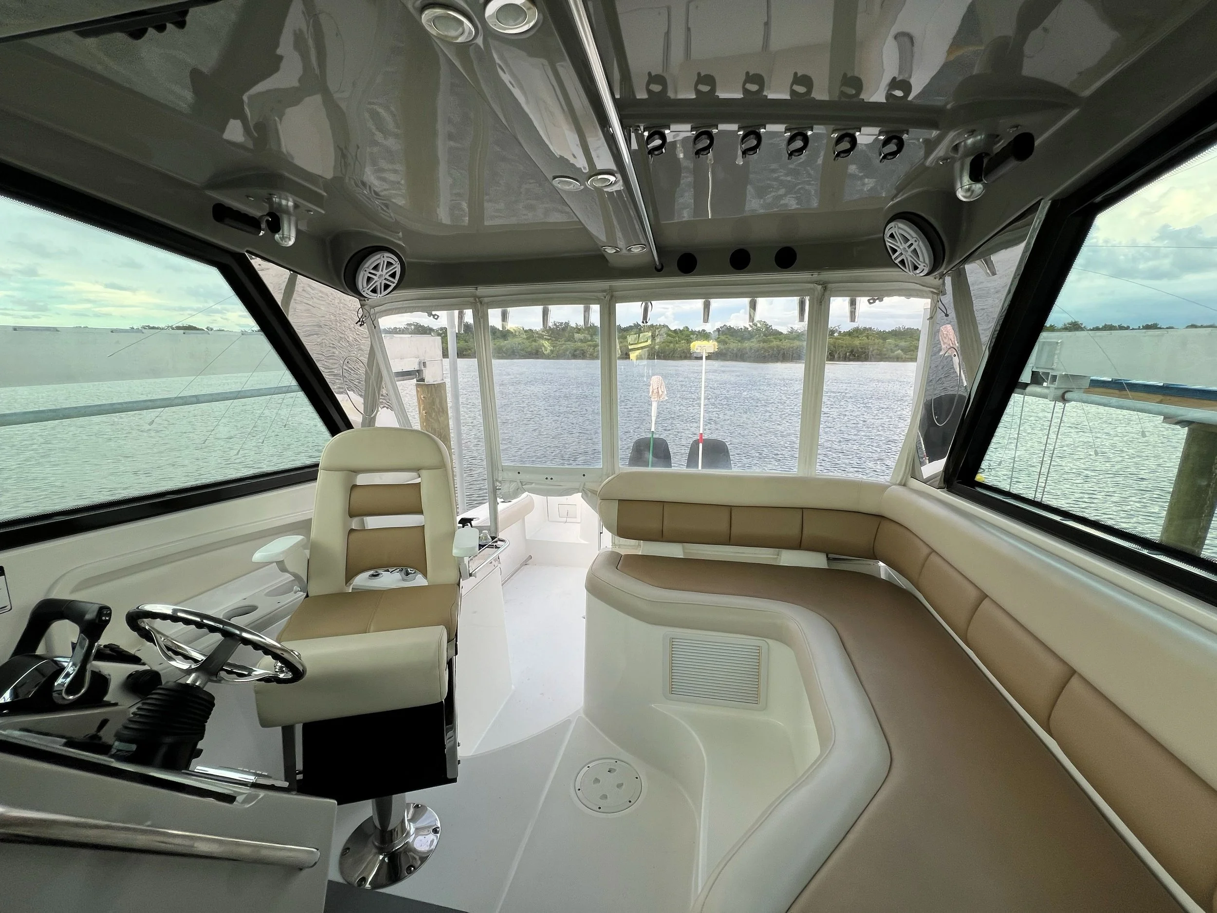 Interior of a boat's cabin with a white captain's chair, beige cushioned seating along the sides, windows revealing water and land outside, and a gray ceiling with ventilation and speaker systems.
