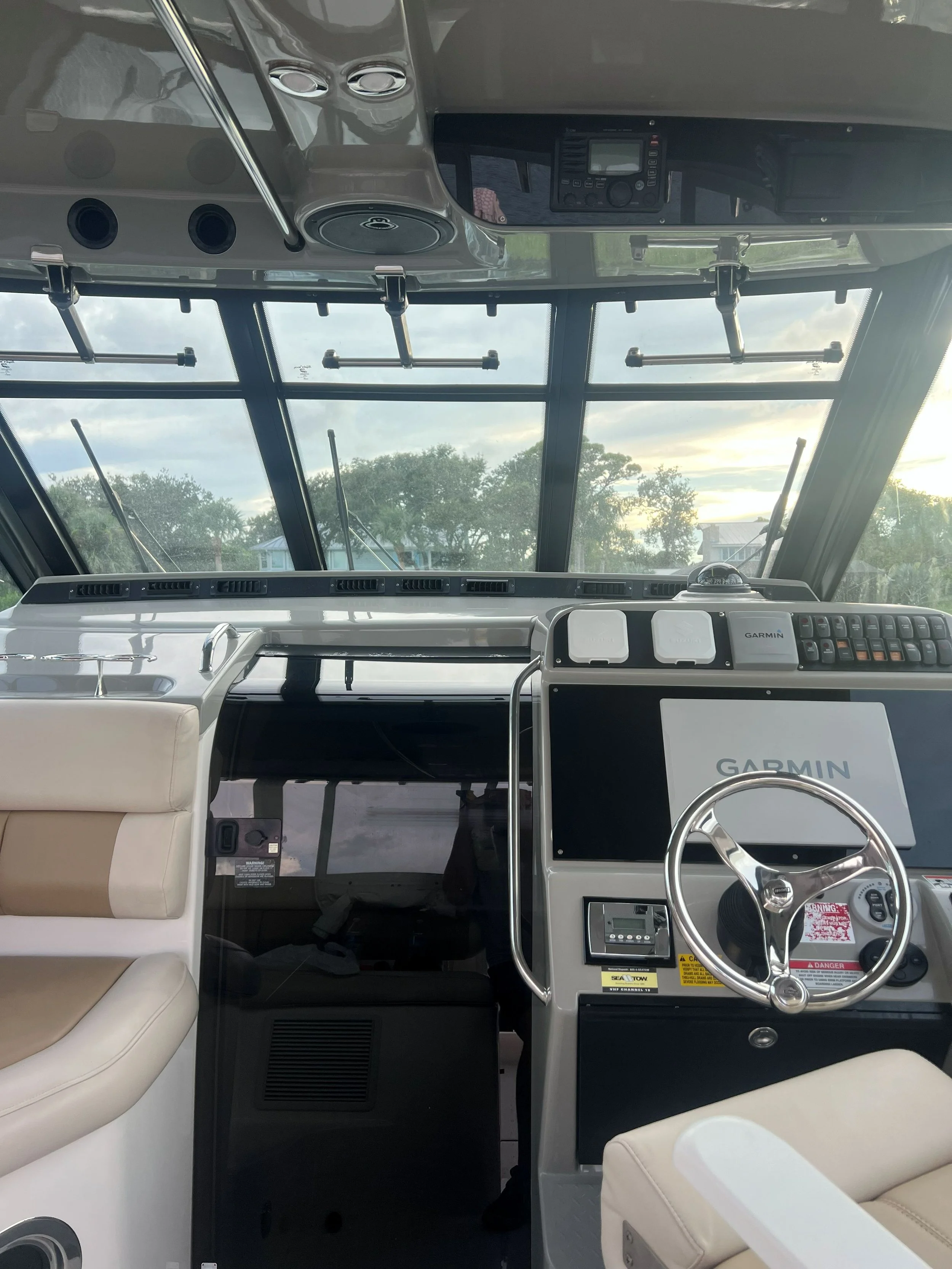 The interior of a boat's cockpit featuring a steering wheel, Garmin navigation screens, control panels, beige seats, and large windows showing trees and sky outside.