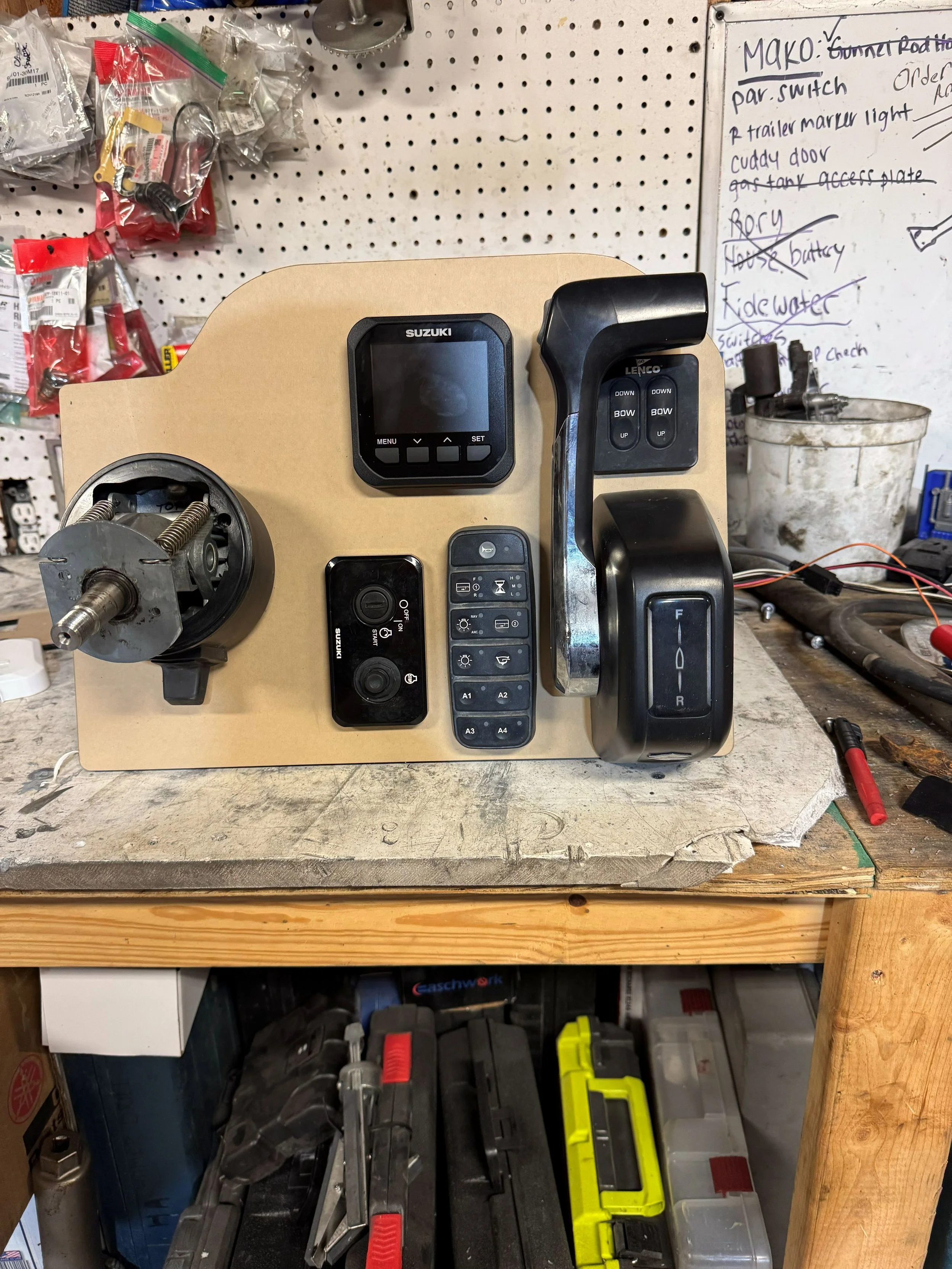Workshop workbench with control panel, joystick, and electronic devices, surrounded by tools, parts, and a pegboard with notes.