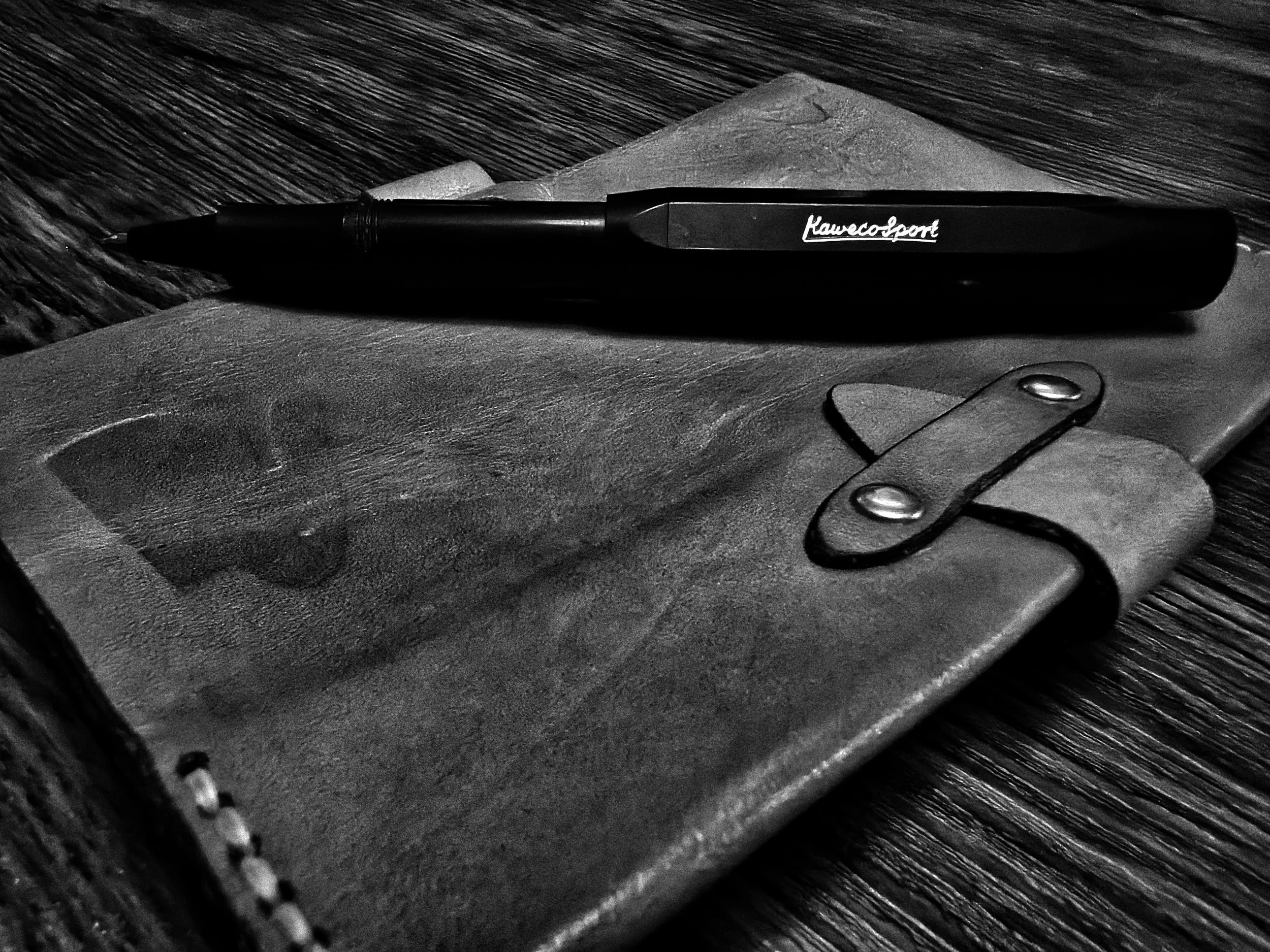 Black and white photo of a leather notebook and a black pen on a wooden surface.