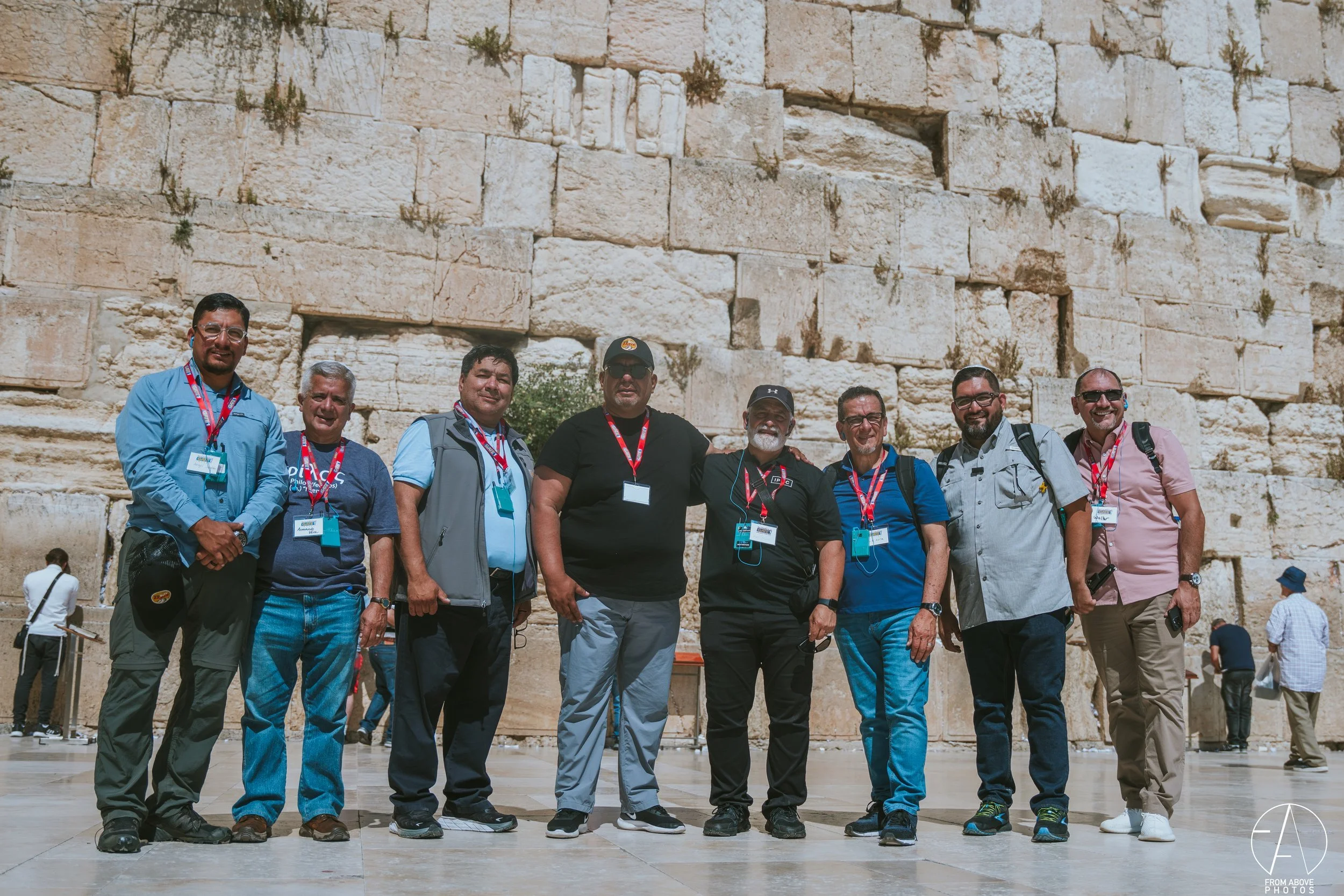 A group of nine men standing together outdoors in front of a large stone wall with small plants growing between the rocks, some of them are wearing sunglasses and all are wearing lanyards with badges.