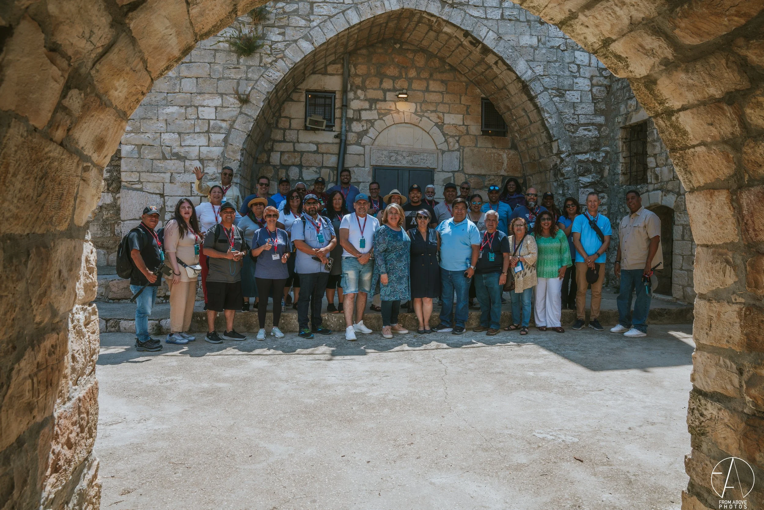 Large group of diverse people posing for a photo in front of an arched stone building, some wearing lanyards, under a stone archway.