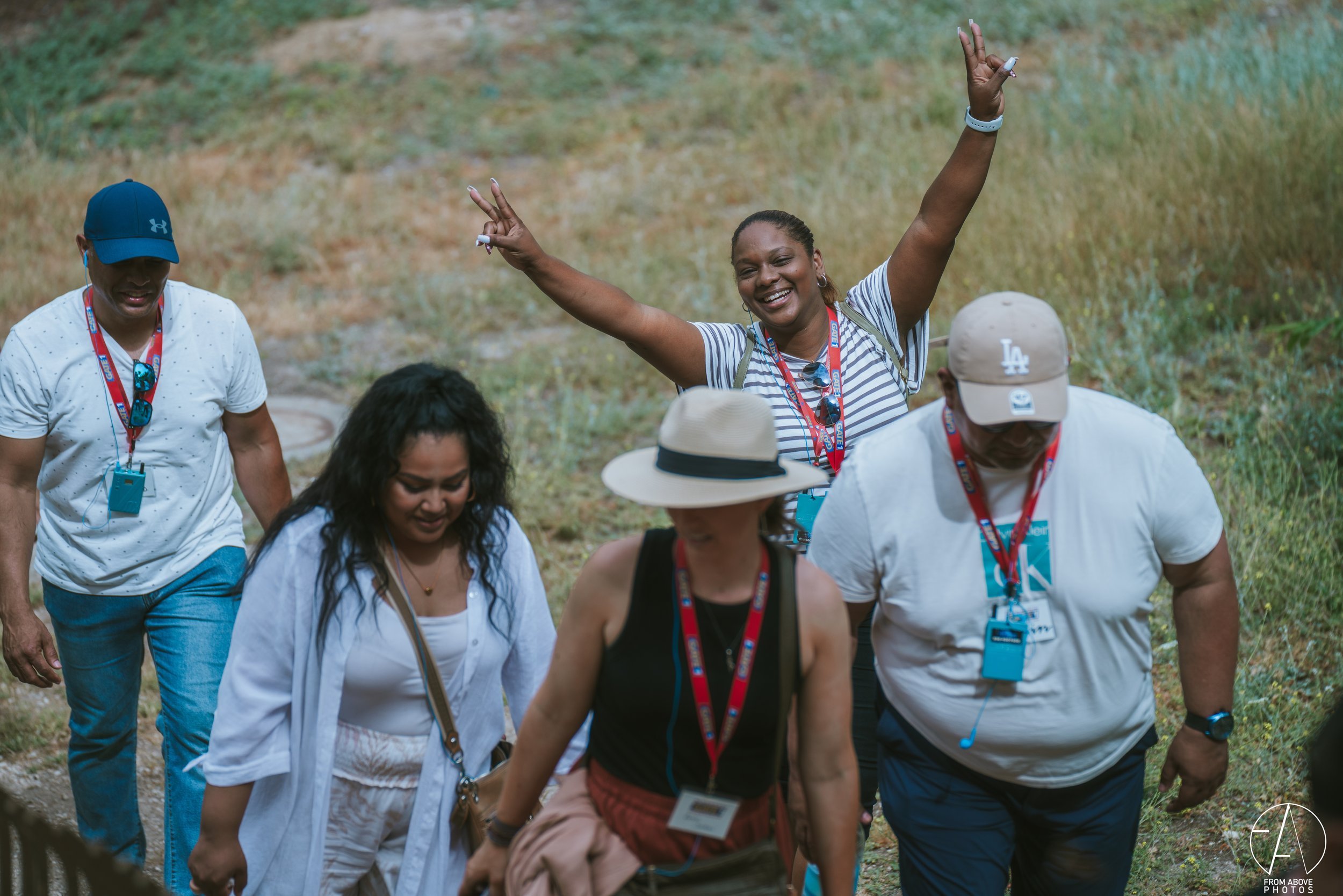 A group of people hiking outdoors on a trail in nature. The woman in the center is smiling with arms raised, wearing a striped shirt and sunglasses on her head. Others around her are looking down or walking, dressed casually, with some wearing hats a