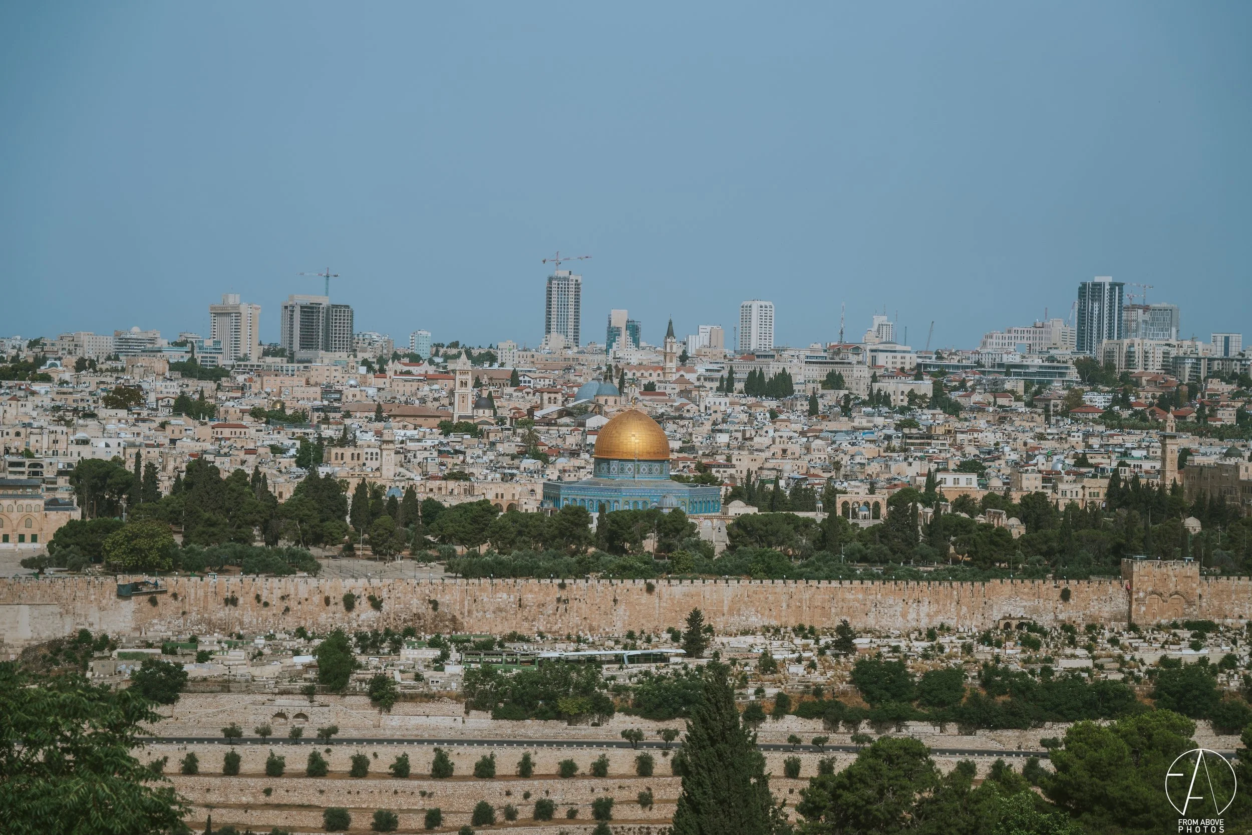 Panoramic view of Jerusalem cityscape with the golden Dome of the Rock in the foreground and modern high-rise buildings in the background under a blue sky.