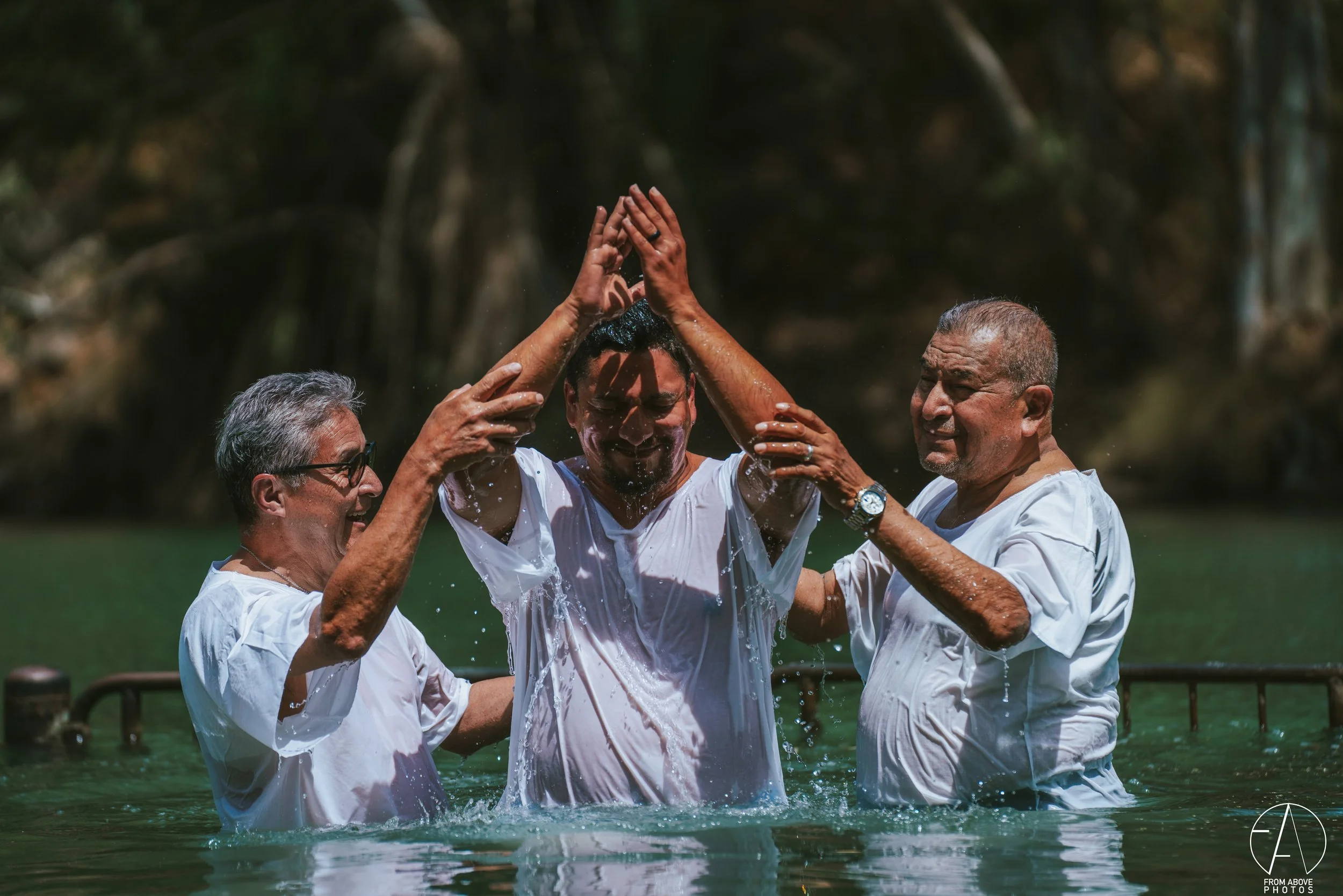 Three men are standing in water outdoors, participating in a baptism ceremony. The man in the center is being baptized, with water pouring over his head as he joins his hands above his head. Two men on either side are supporting and assisting with th