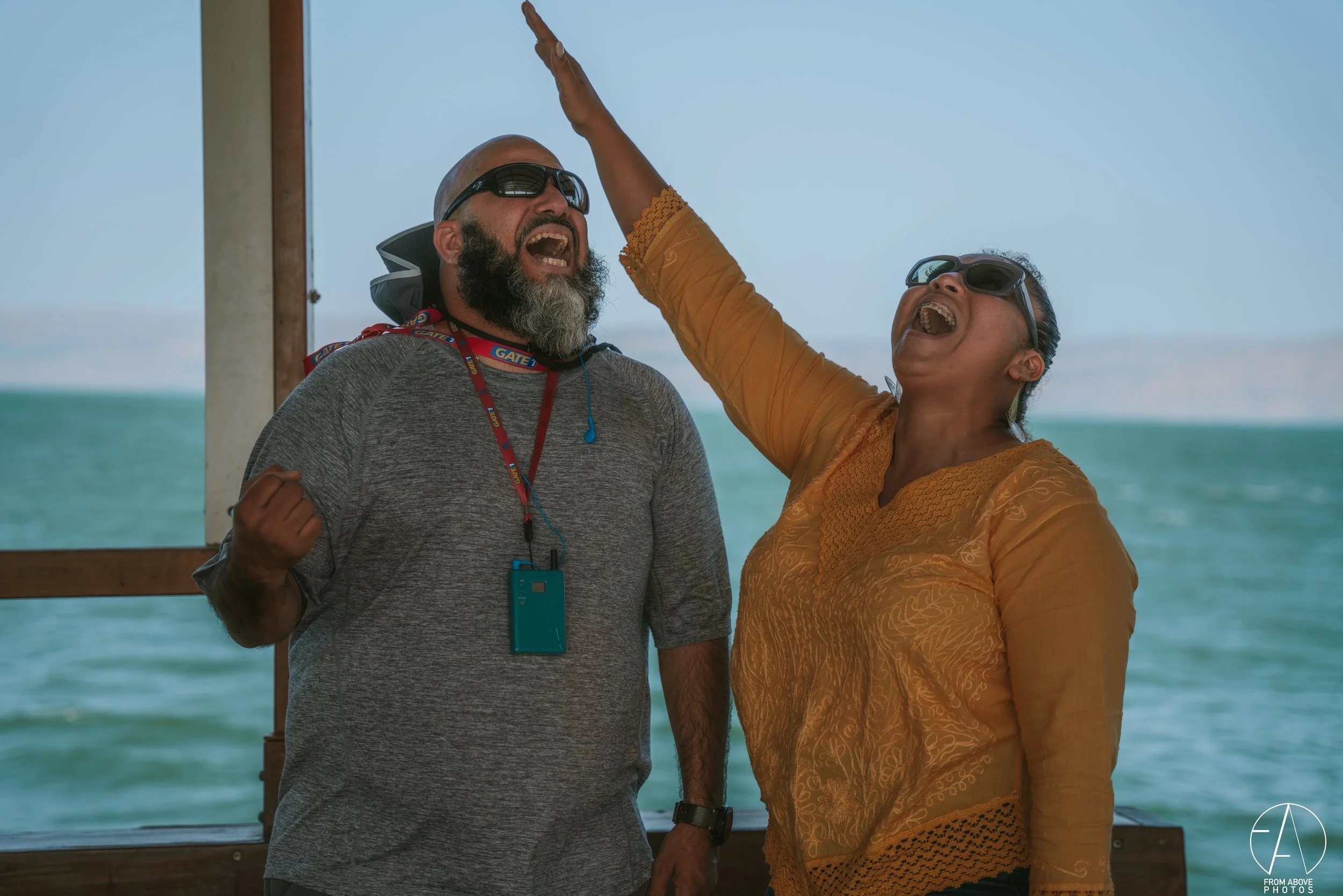 A man and a woman are celebrating on a boat, wearing sunglasses, with water in the background. The man has a beard and is wearing a grey shirt, and the woman in a yellow blouse. They are smiling and laughing, appearing joyful.