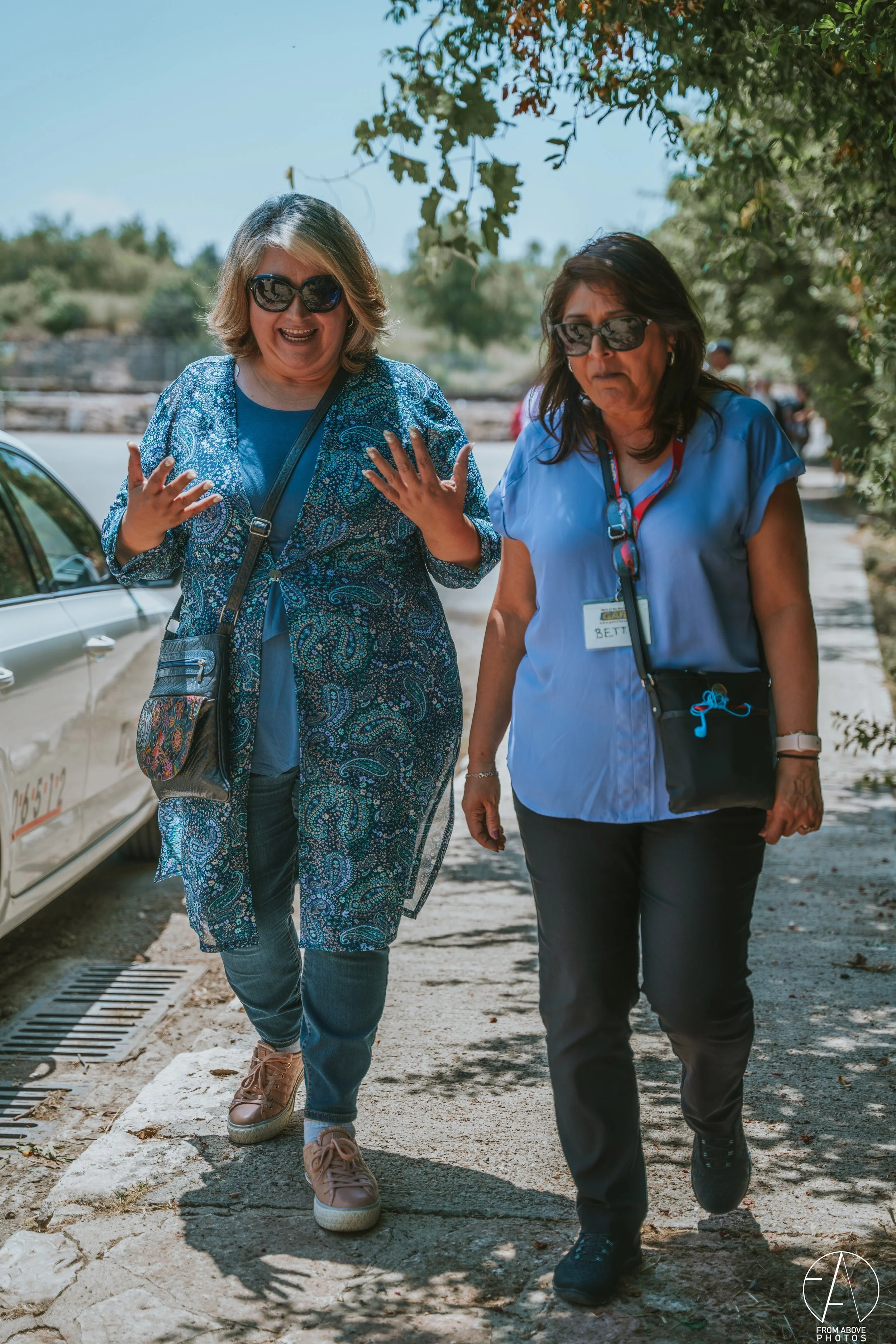 Two women walking outdoors on a sunny day, one wearing sunglasses and a blue patterned top, the other also with sunglasses and a blue blouse, surrounded by trees and parked cars.