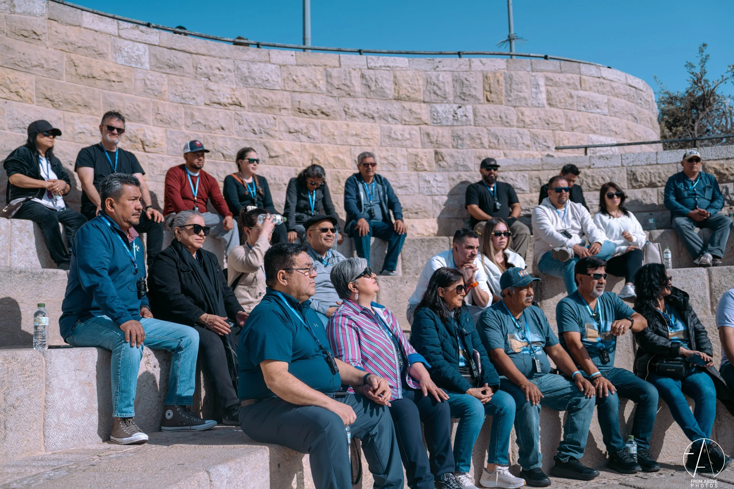 A group of people sitting and standing on stone steps outdoors, attending an event on a sunny day.