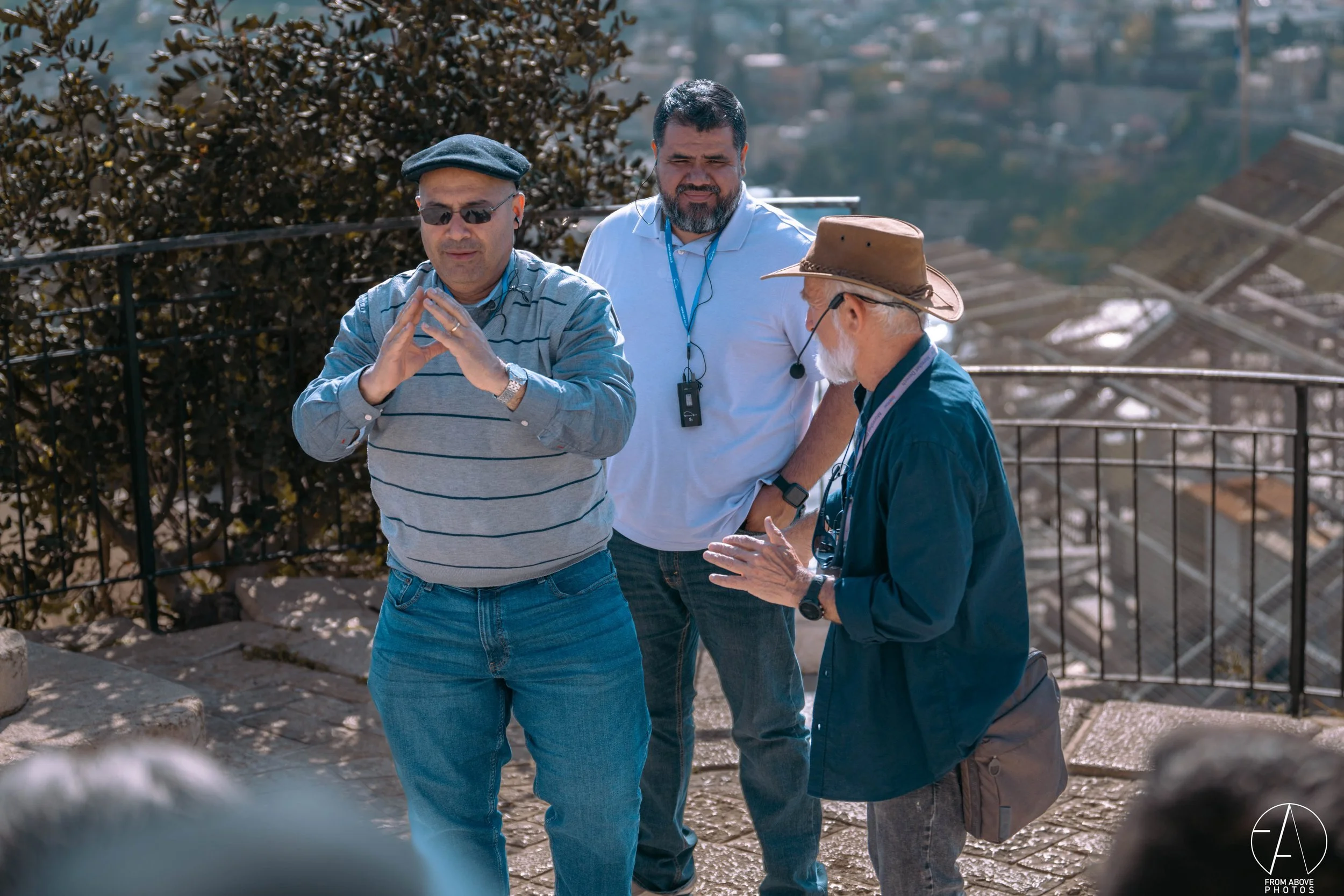 Three men standing outdoors on a sunny day, engaged in a conversation. One man is gesturing with his hands, wearing sunglasses, a gray striped sweater, and a flat cap. The second man, dressed in a white shirt with a lanyard around his neck, is listen