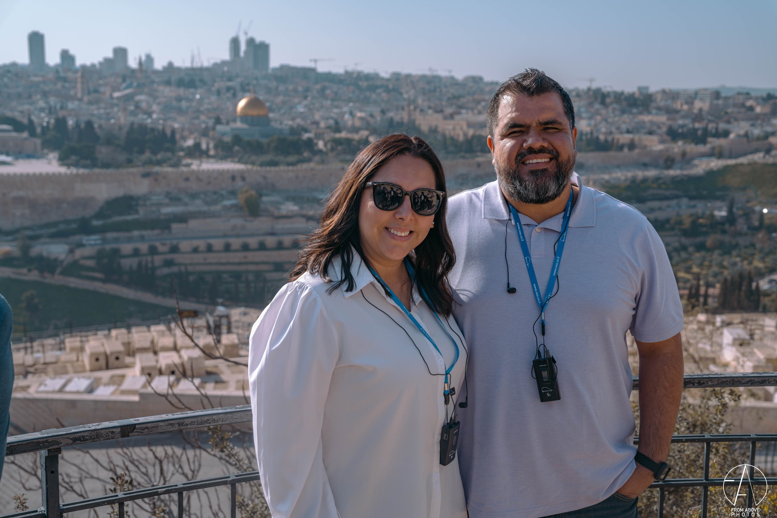 A woman wearing sunglasses and a white blouse and a man wearing a light gray polo shirt with a beard, both with walkie-talkies around their necks, standing on a viewing platform with the city skyline and the Dome of the Rock in Jerusalem in the backg