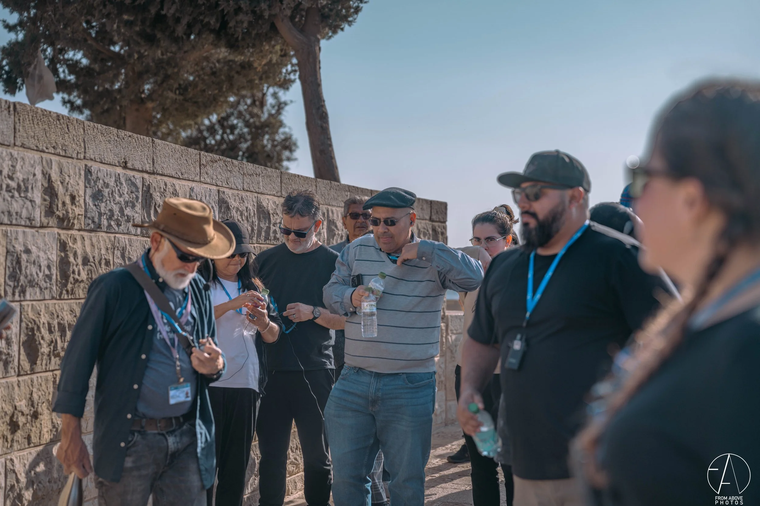 A group of people standing outdoors near a stone wall, holding water bottles and wearing sunglasses. The group appears to be engaged in a discussion or listening to a guide, with some wearing ID badges and lanyards. The scene is sunny with trees in t
