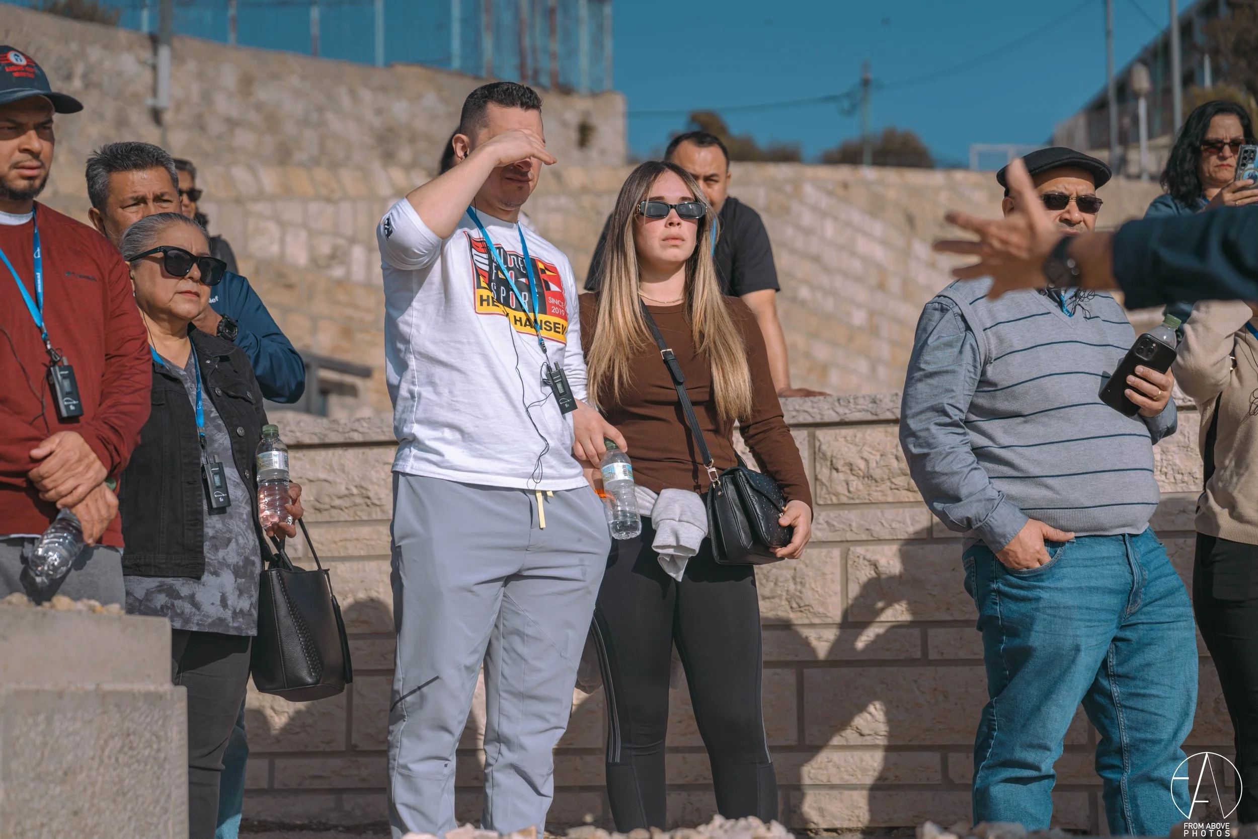 Group of people standing outdoors, some wearing sunglasses, holding water bottles, and listening to a person gesturing with their hand.