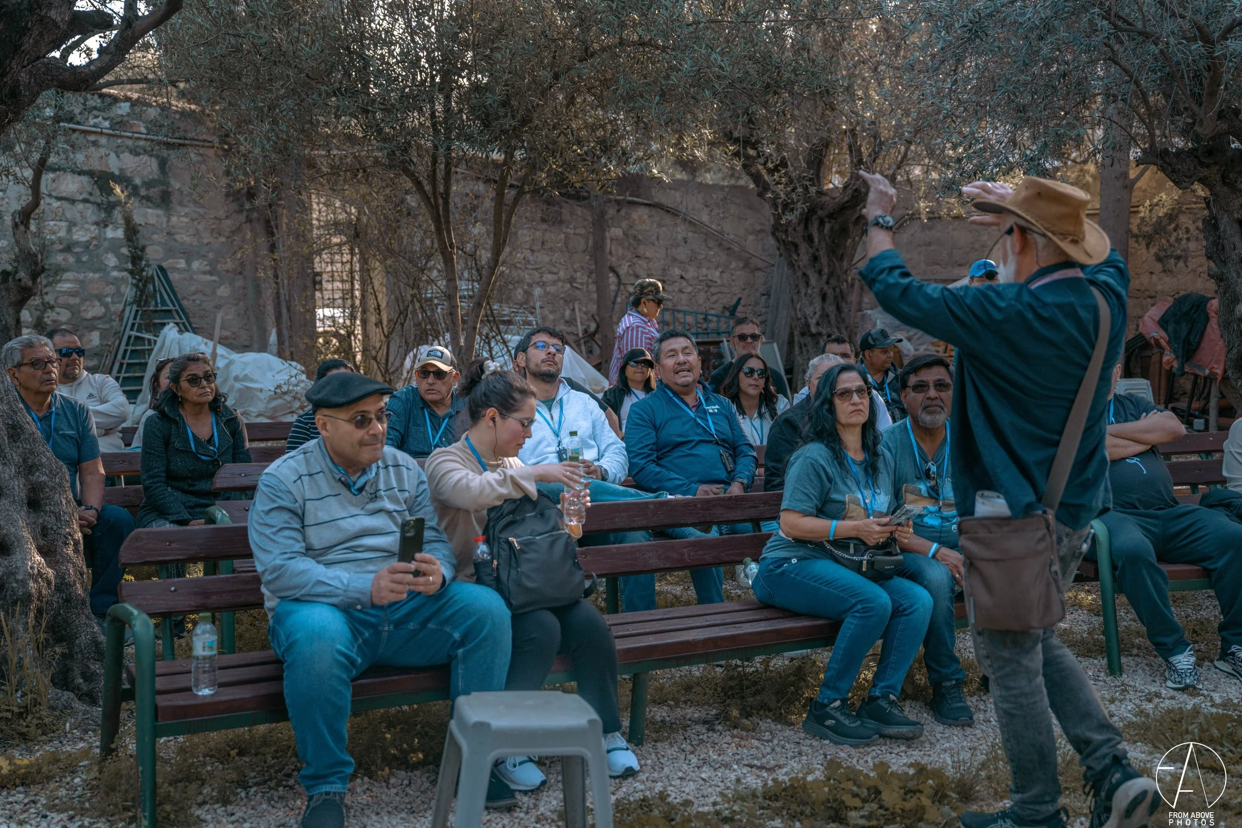 A group of people seated on benches outdoors listening to a man standing in front of them, gesturing with his hand. The setting appears to be a park or garden with trees and a stone wall in the background.