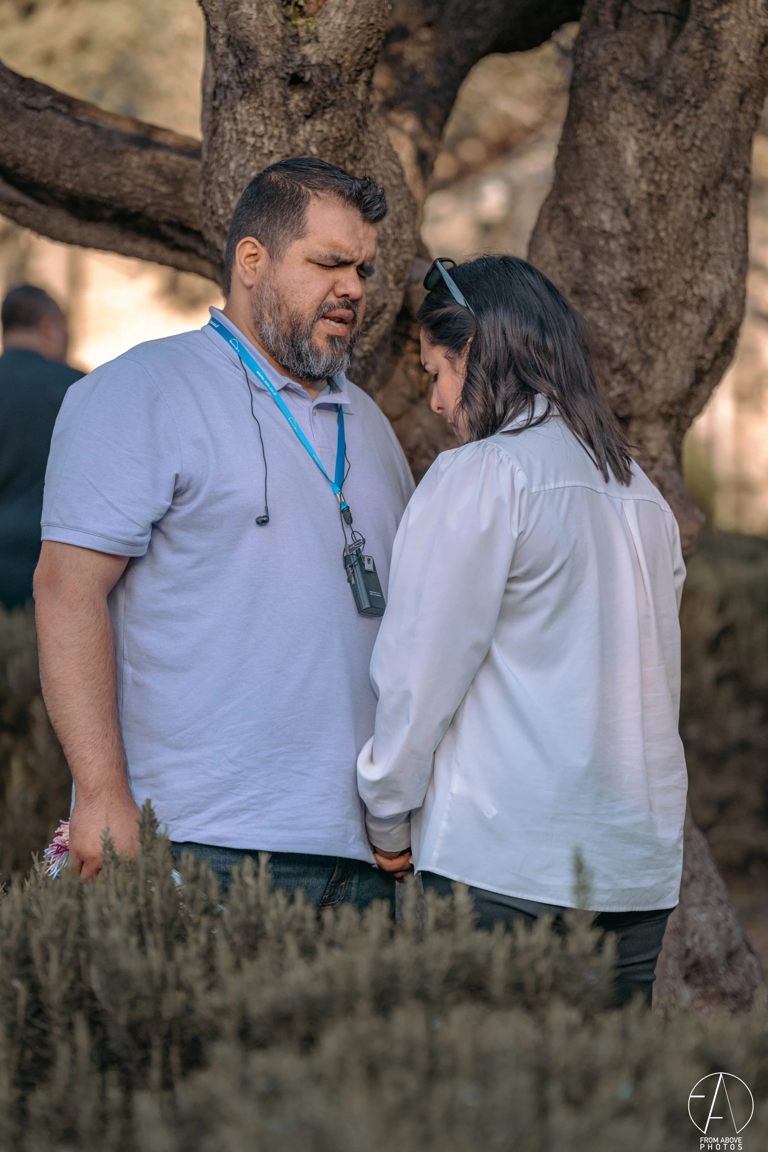 Two people praying with heads bowed, standing outdoors near a large tree, holding hands, showing a moment of reflection or prayer.