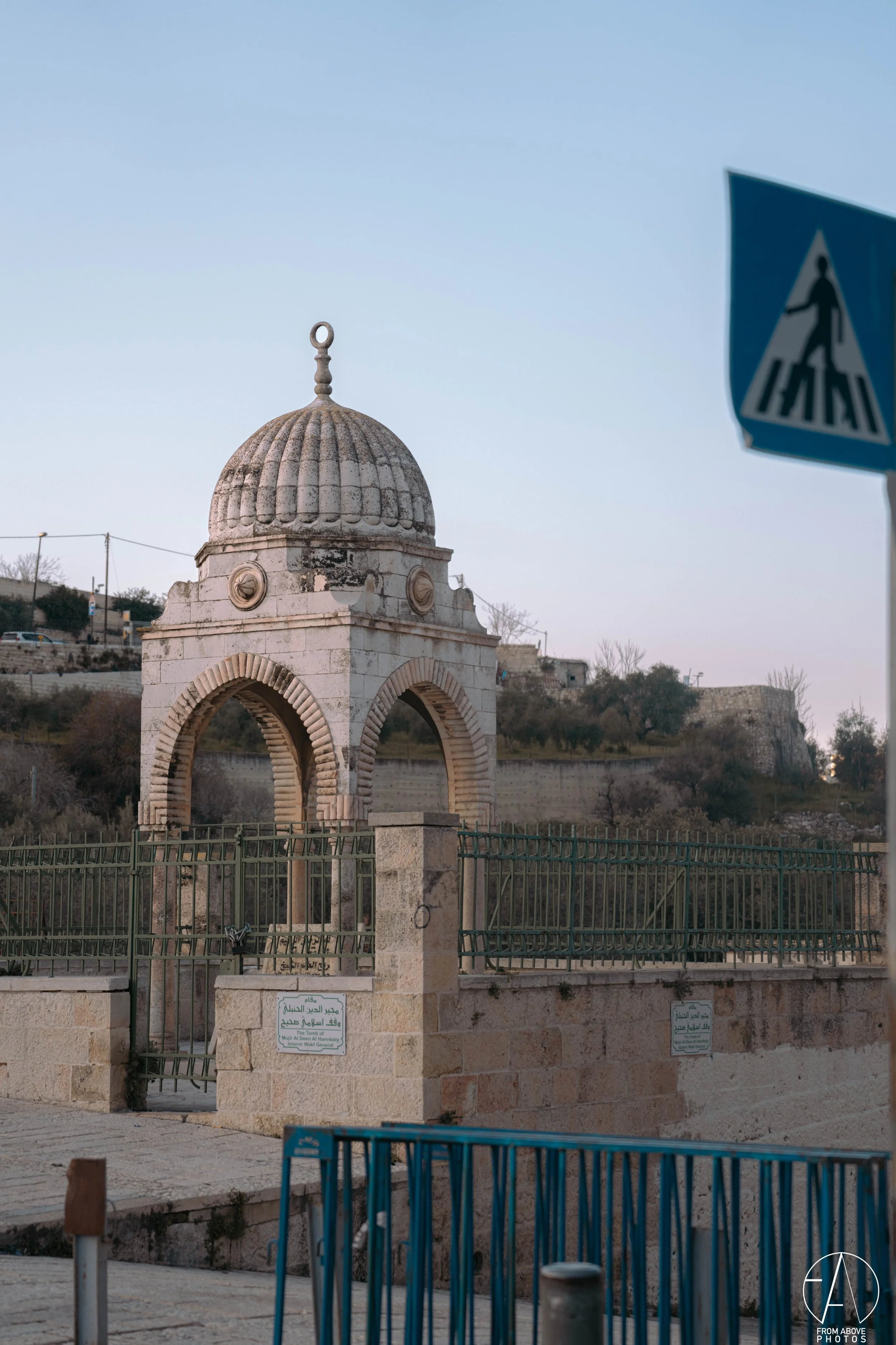 A small stone pavilion with a domed roof and arched openings, fenced in with green metal fencing, situated on a stone platform with a blue pedestrian crossing sign in the foreground, under a cloudy sky.