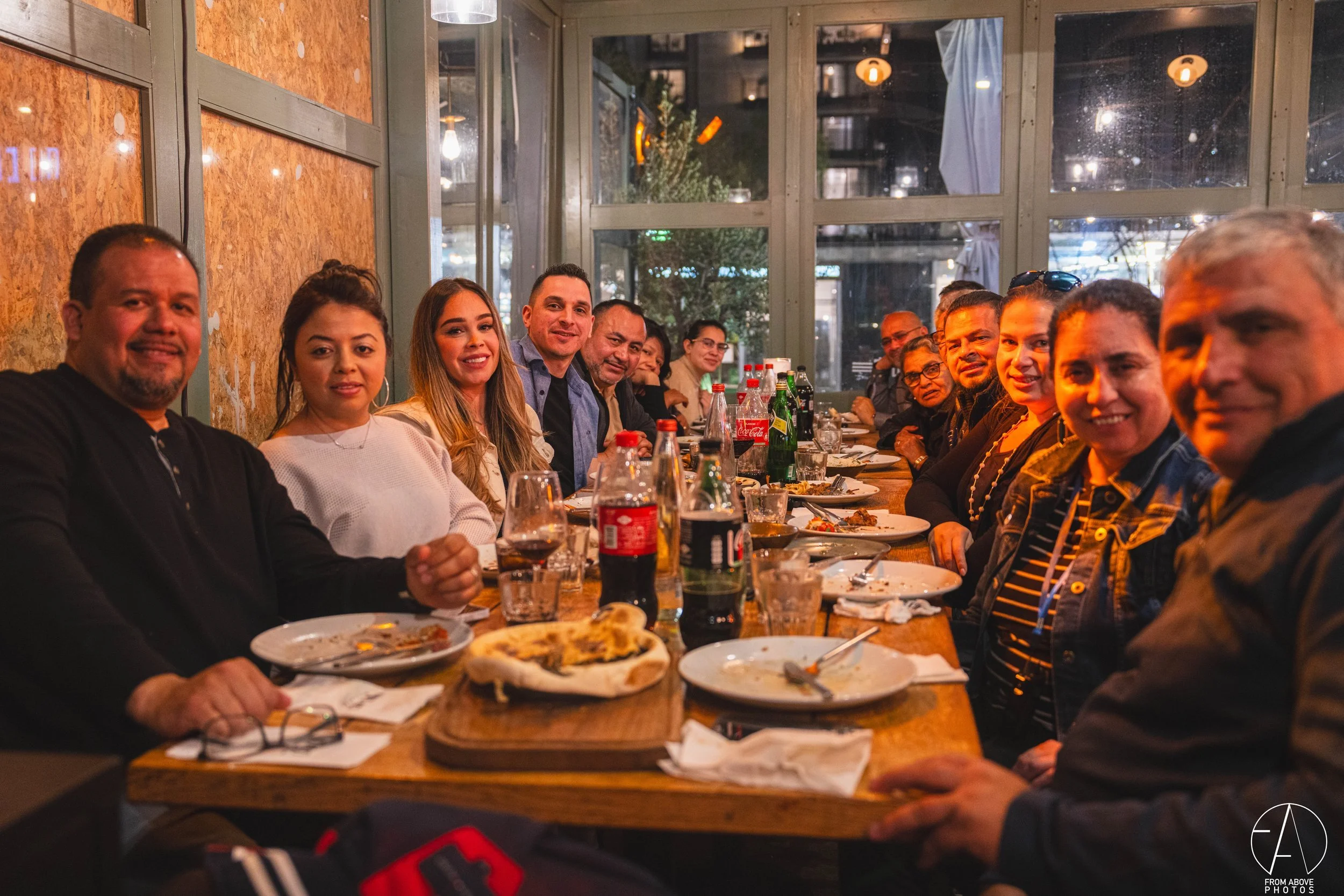A group of people sitting at a long table in a restaurant, smiling and enjoying a meal together with empty plates, glasses, and bottles on the table.