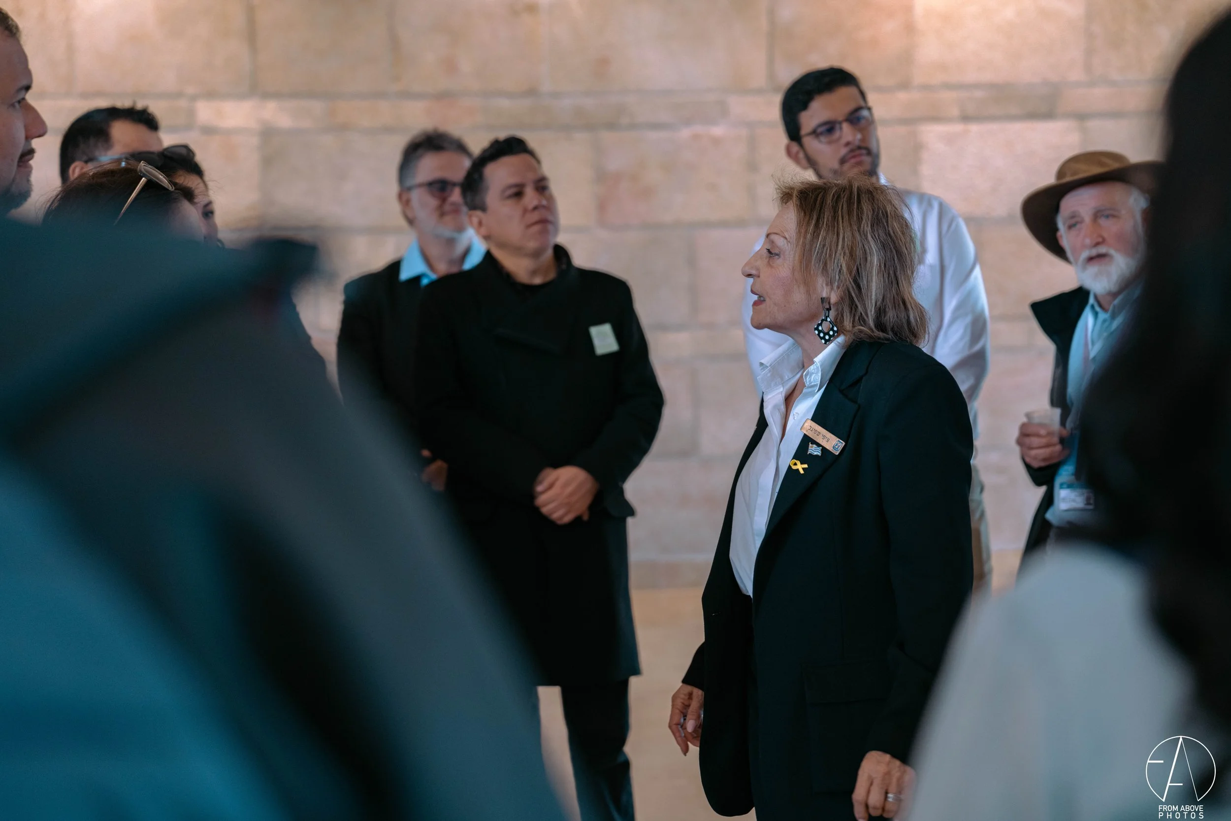 A woman in a black blazer and white shirt speaking to a group of people indoors, with a beige brick wall in the background.