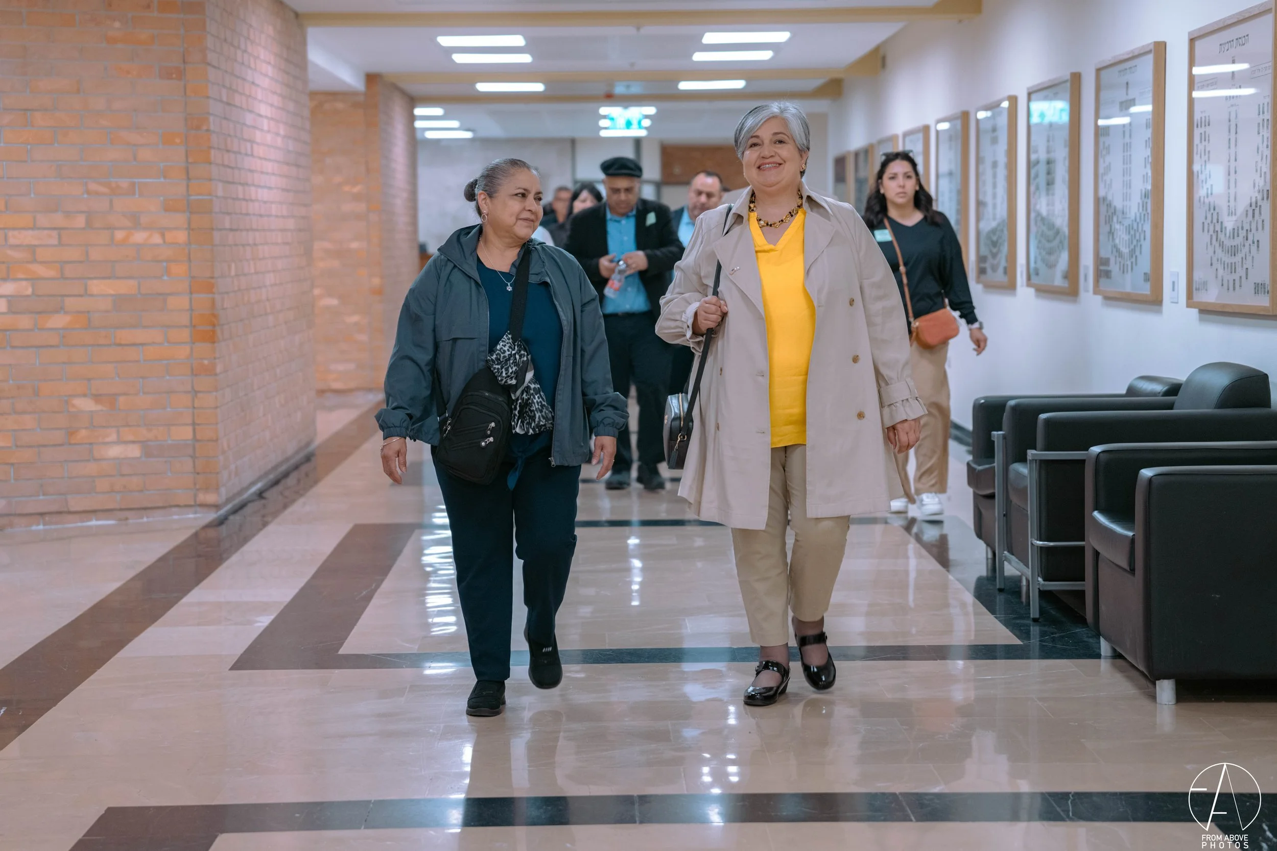 A group of people walking down a hallway in a building, including an older woman in a beige coat and yellow top smiling, with others following behind.