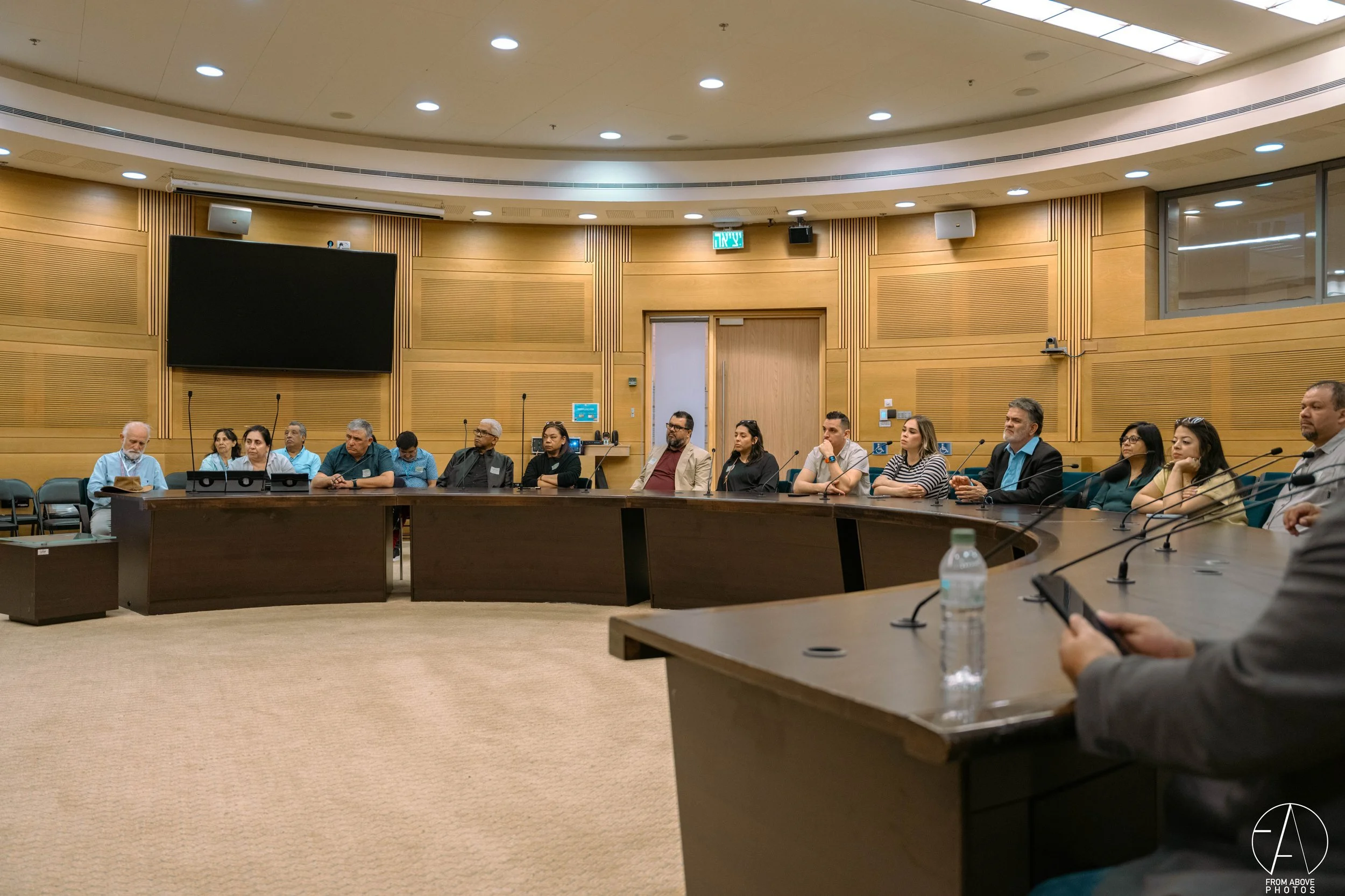 A group of people seated around a large, U-shaped conference table in a wood-paneled room with microphones, a water bottle, and a large television screen on the wall.