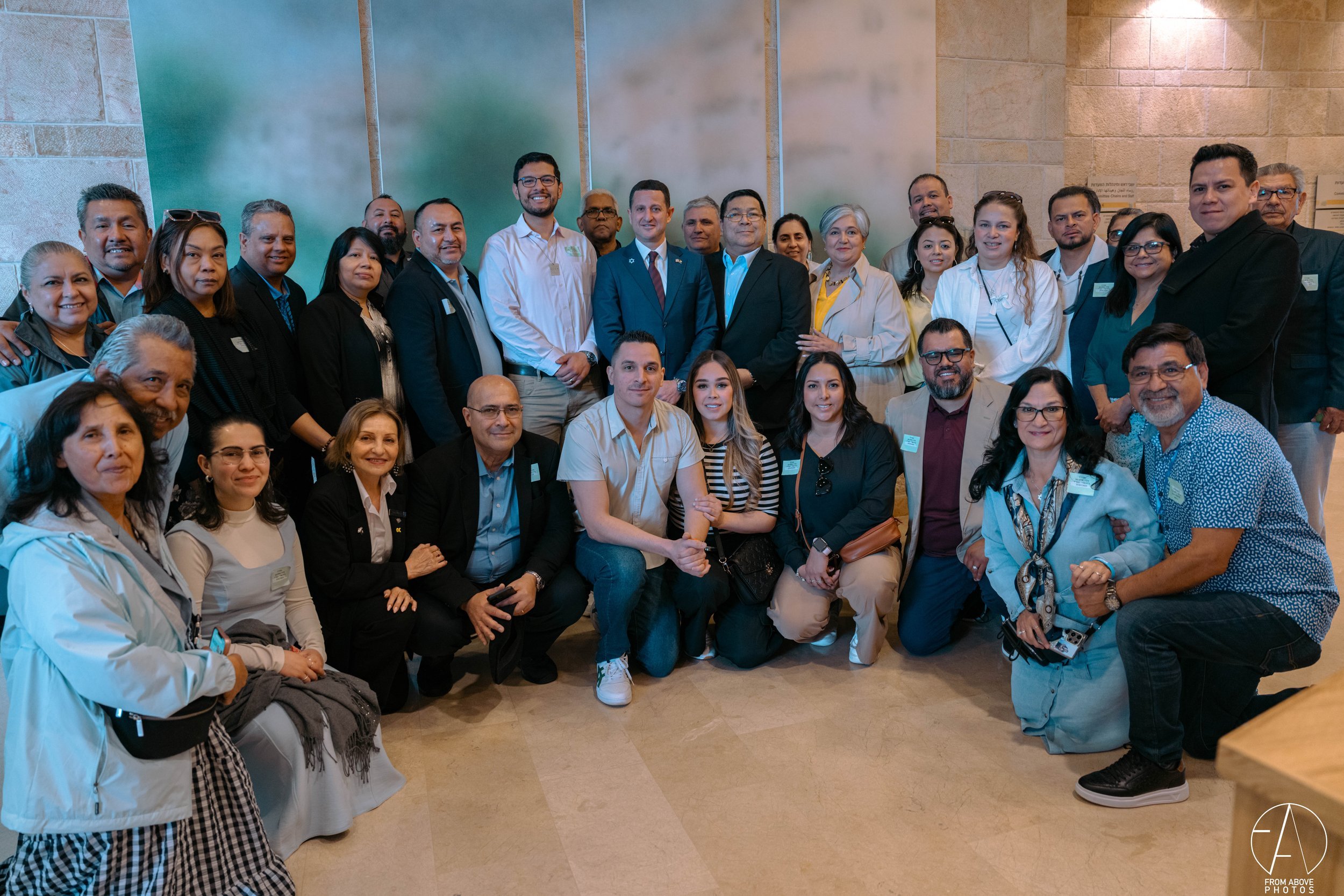 A large group of people posing for a photo indoors in front of frosted glass and stone walls, some standing and some kneeling or sitting, smiling at the camera.