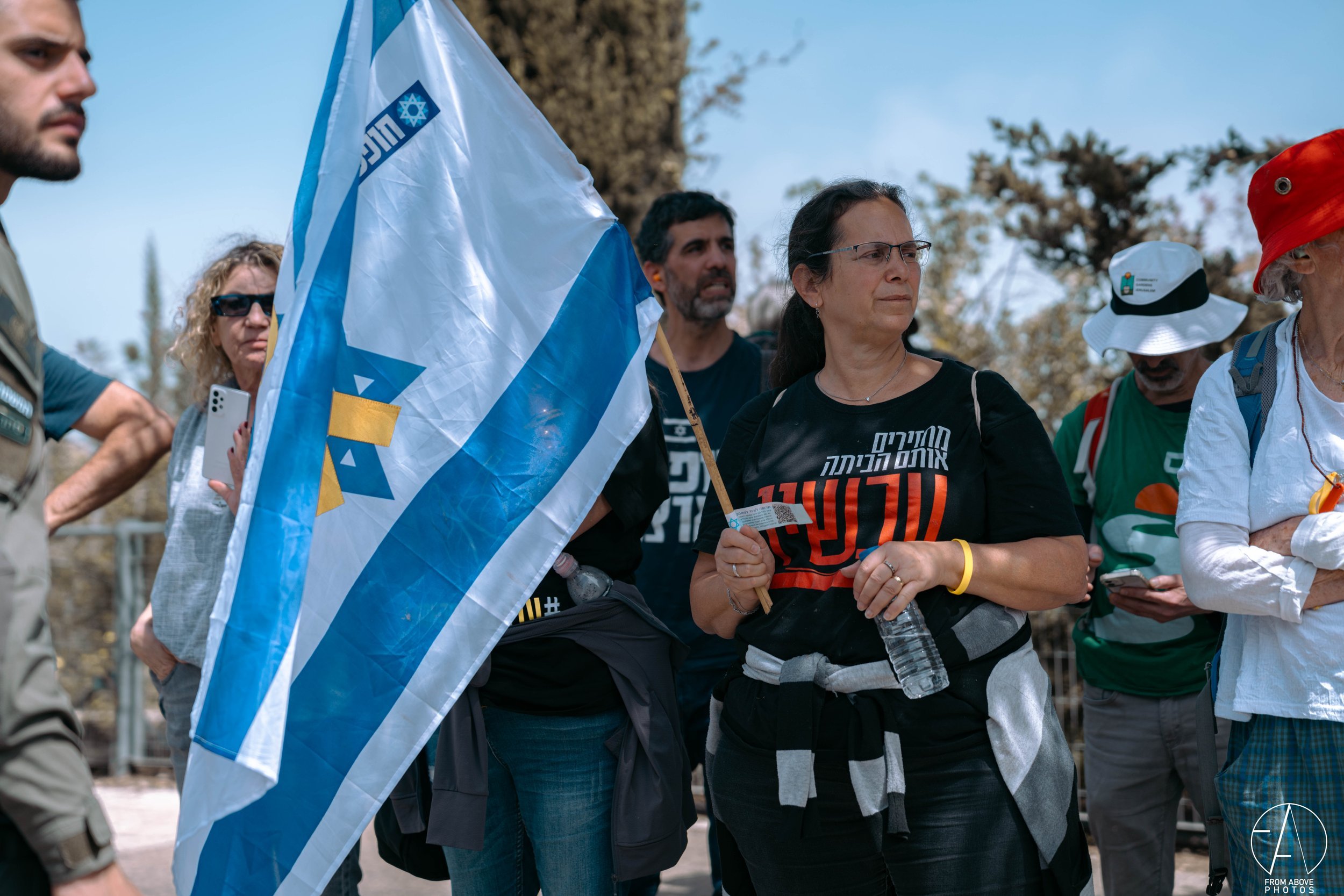 Group of people gathered outdoors, one woman holding an Israeli flag, some using smartphones, dressed casually, with trees and a clear sky in the background.