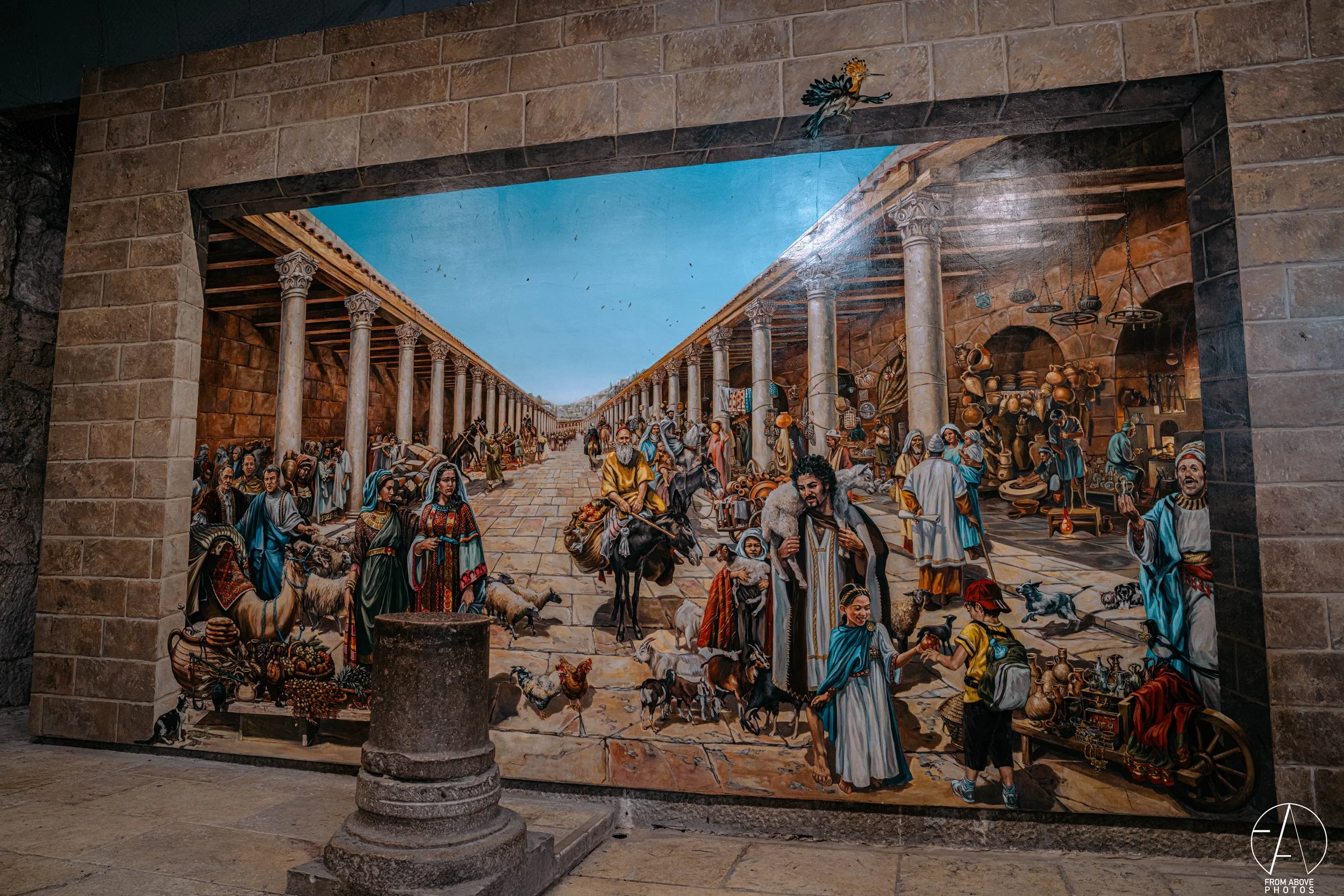 Mural depicting a bustling ancient marketplace with people, animals, and vendors under a covered walkway with columns, against a clear blue sky.