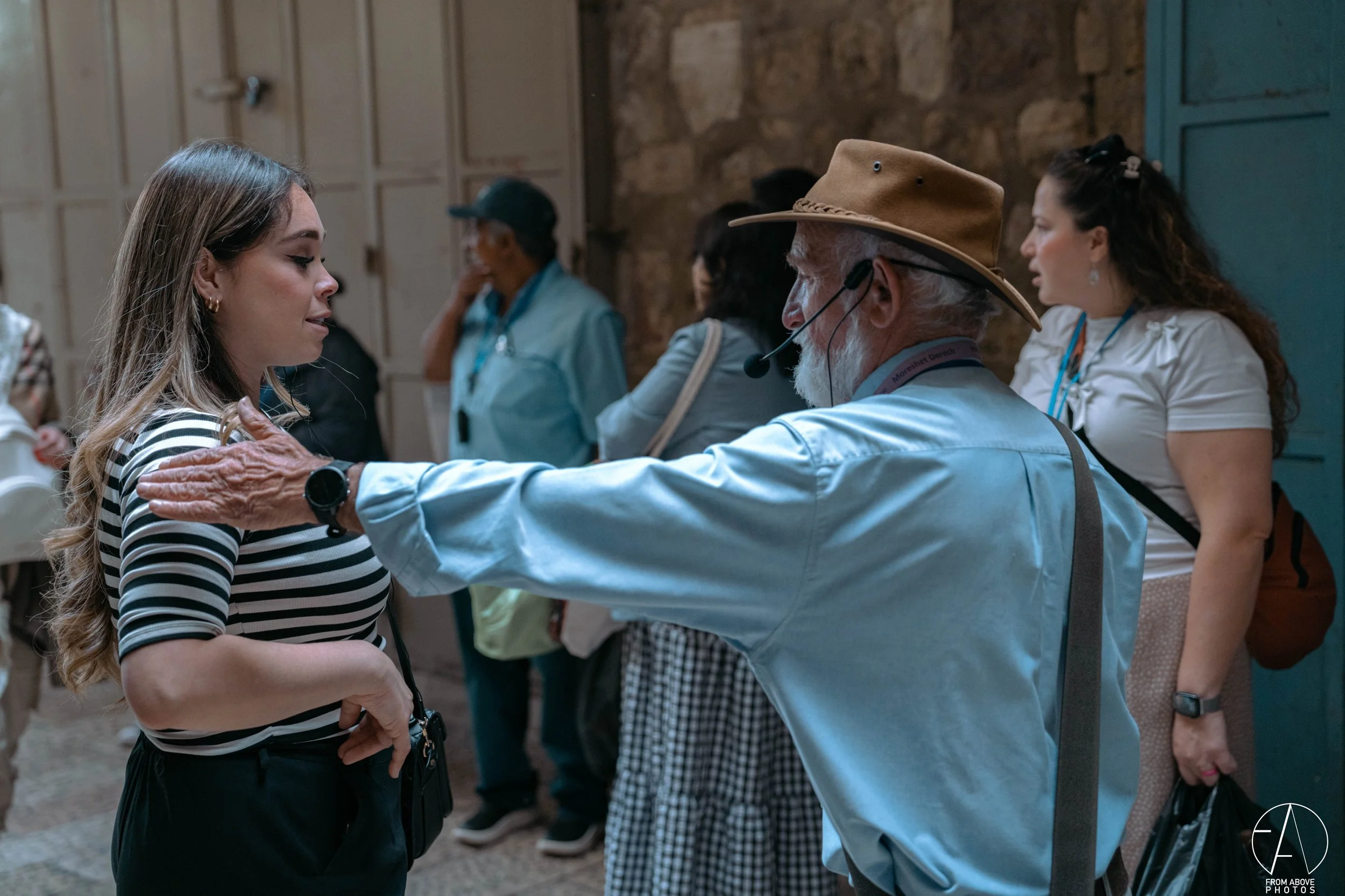 An elderly man with a microphone headset places his hand on a young woman's shoulder in a friendly manner during a discussion or interview, with several people in the background at an indoor event.