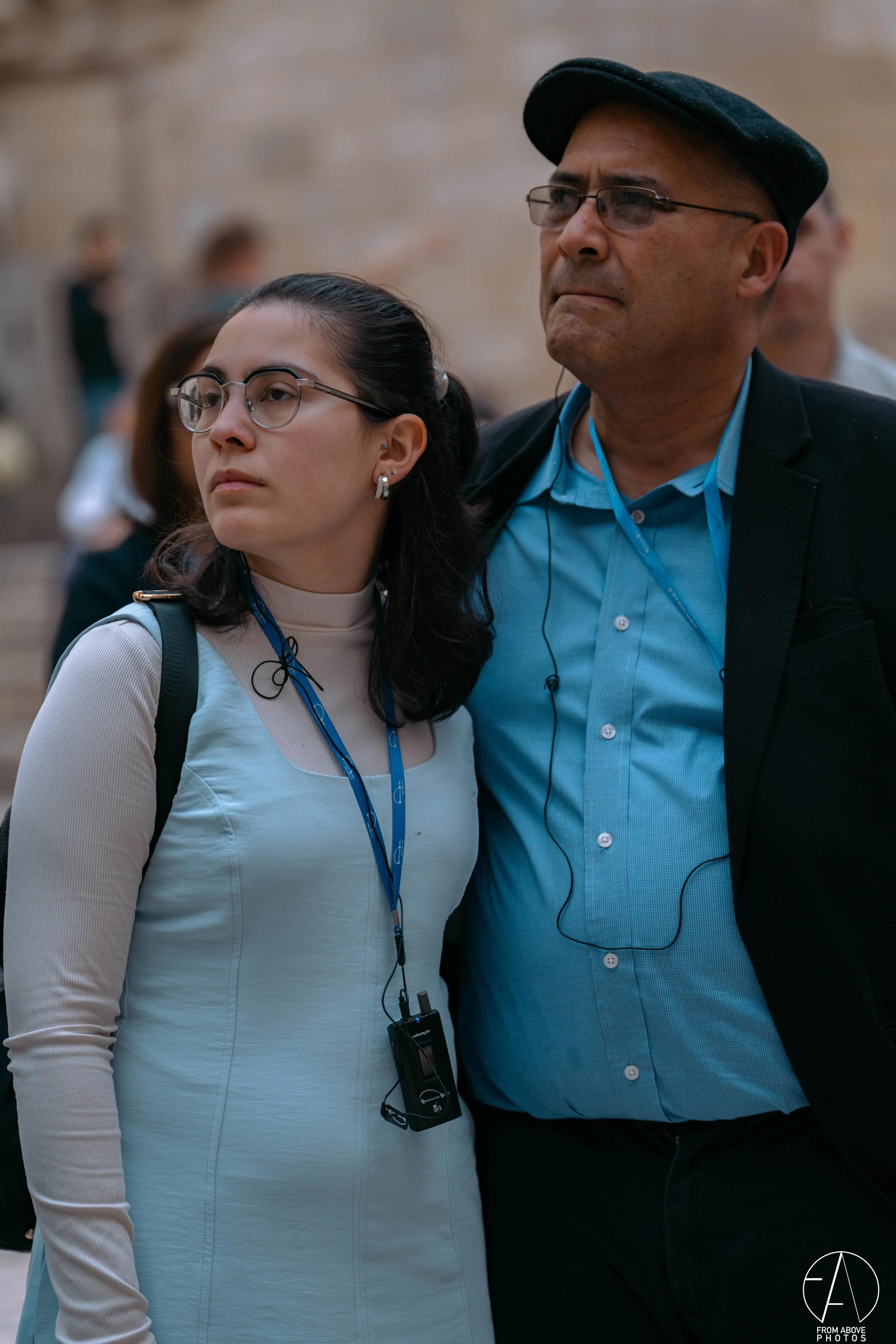 A man and woman standing close together, both wearing glasses and listening to earpieces, in an outdoor setting with blurred people in the background.