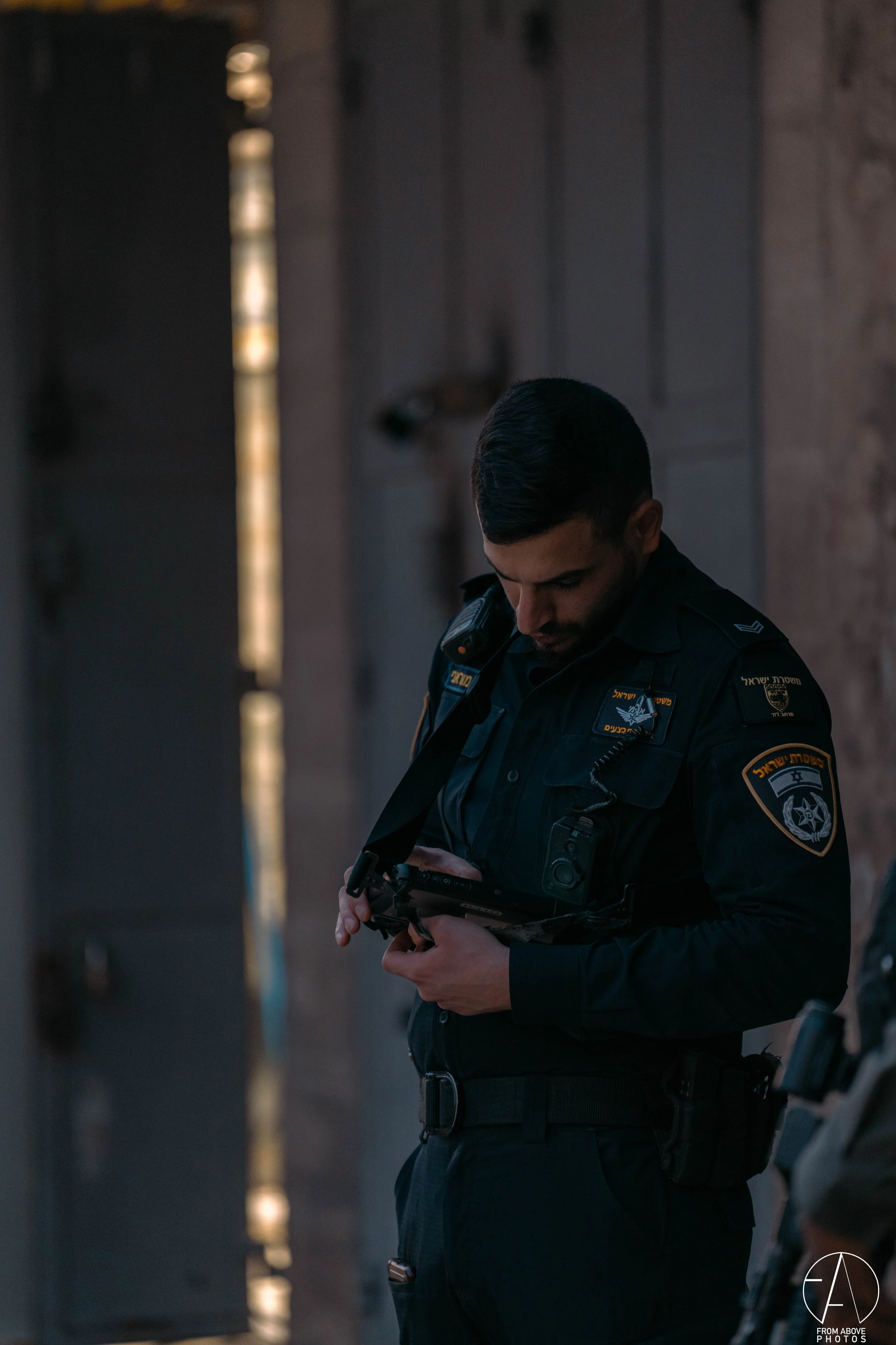A police officer in uniform is looking down at his tablet device inside a room with a wooden wall and a partially open door, with light coming through the door cracks.