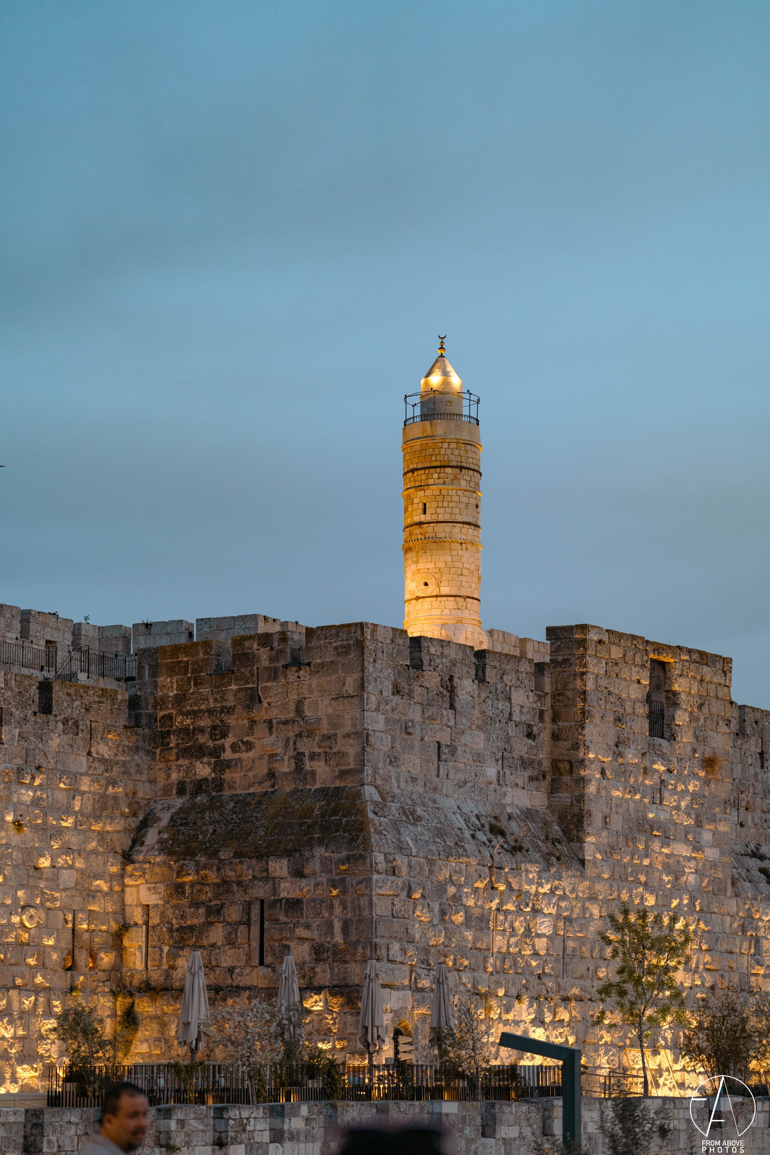 A historic stone fortress wall with a tall lighthouse on top, illuminated at dusk, and umbrellas in the foreground.