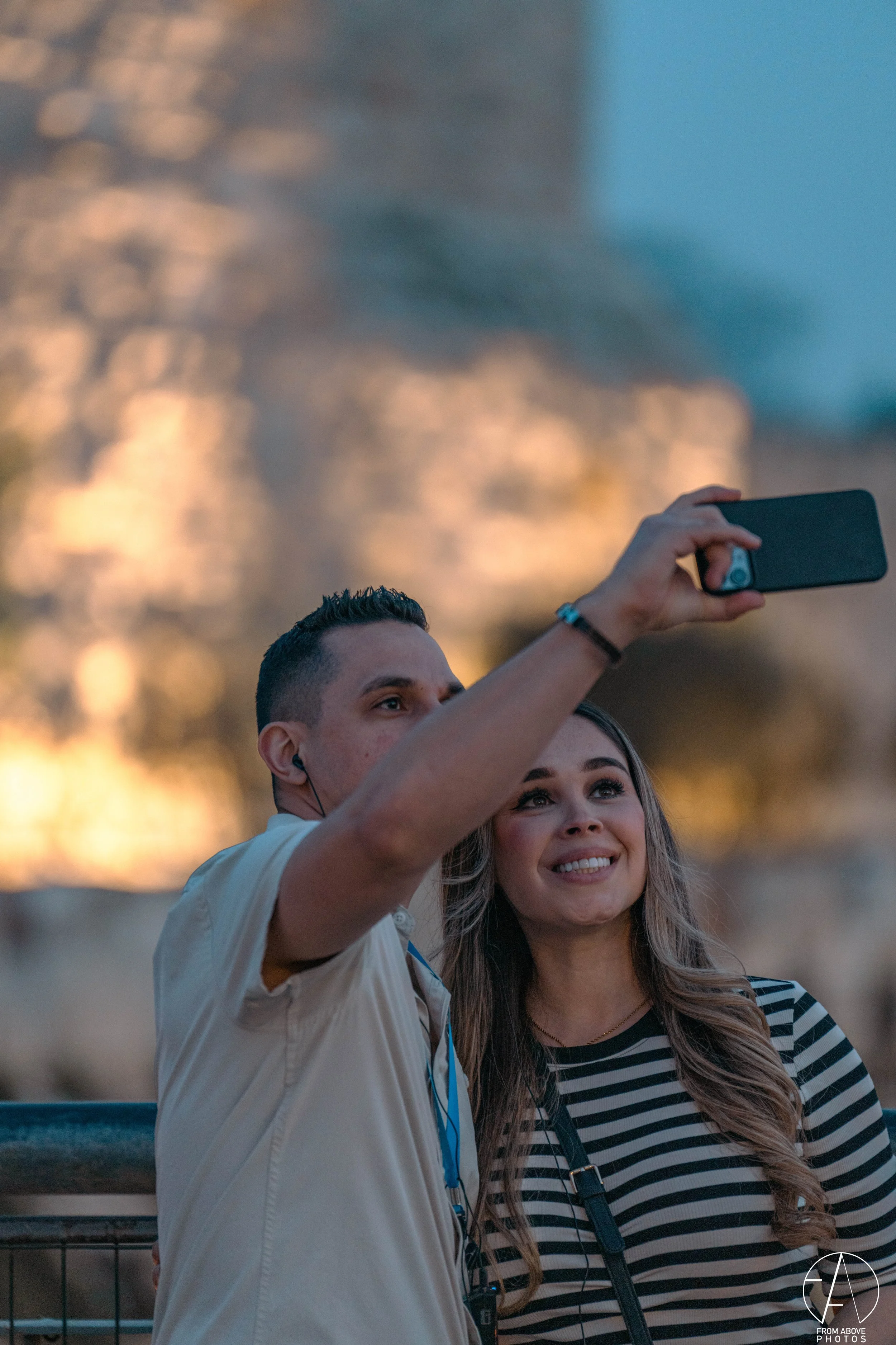A man and a woman taking a selfie together outdoors during sunset. The man is holding a smartphone and the woman is smiling at the camera.