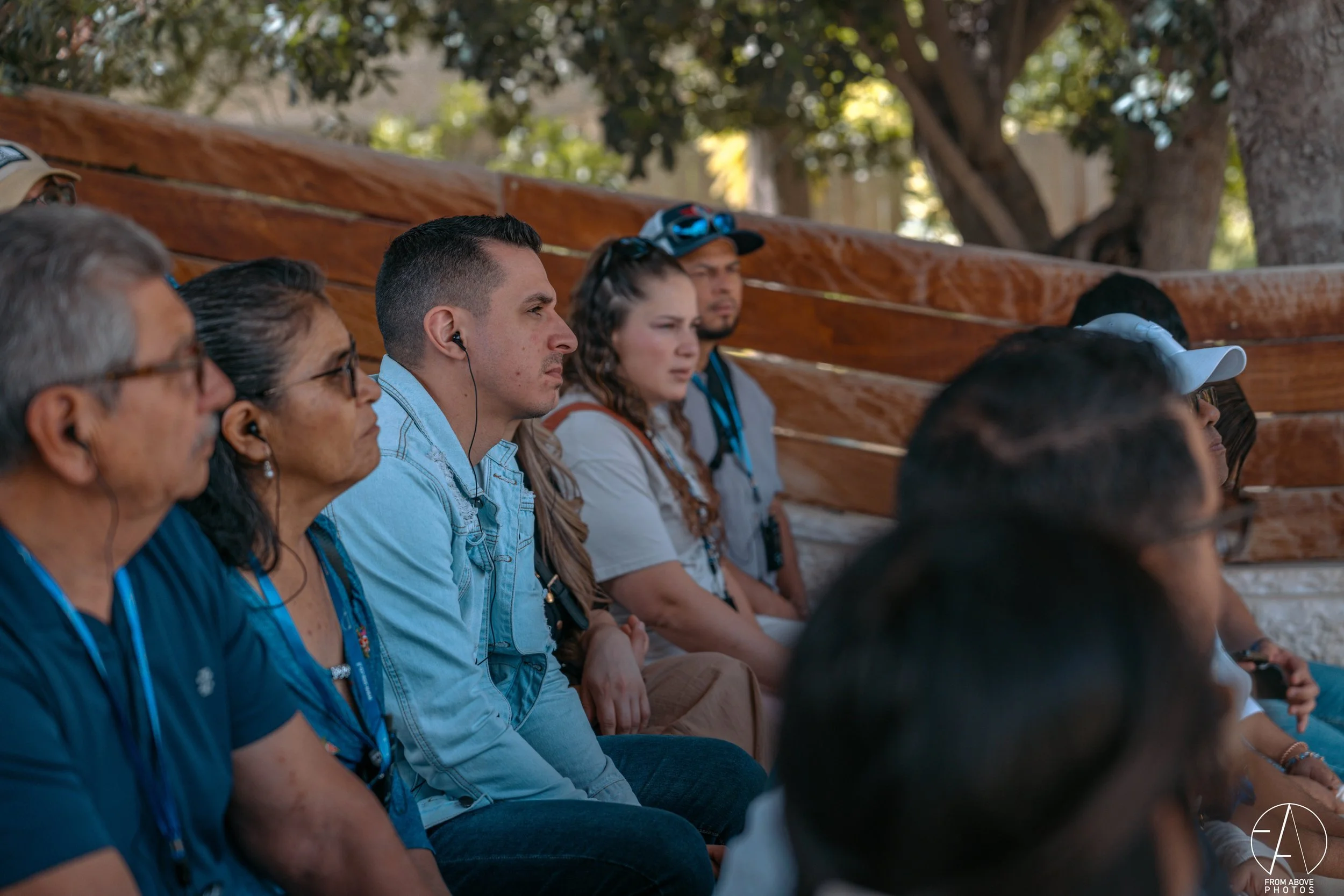 Group of diverse adults sitting outdoors, listening attentively, some wearing headphones and lanyards, with trees and wooden benches in the background.