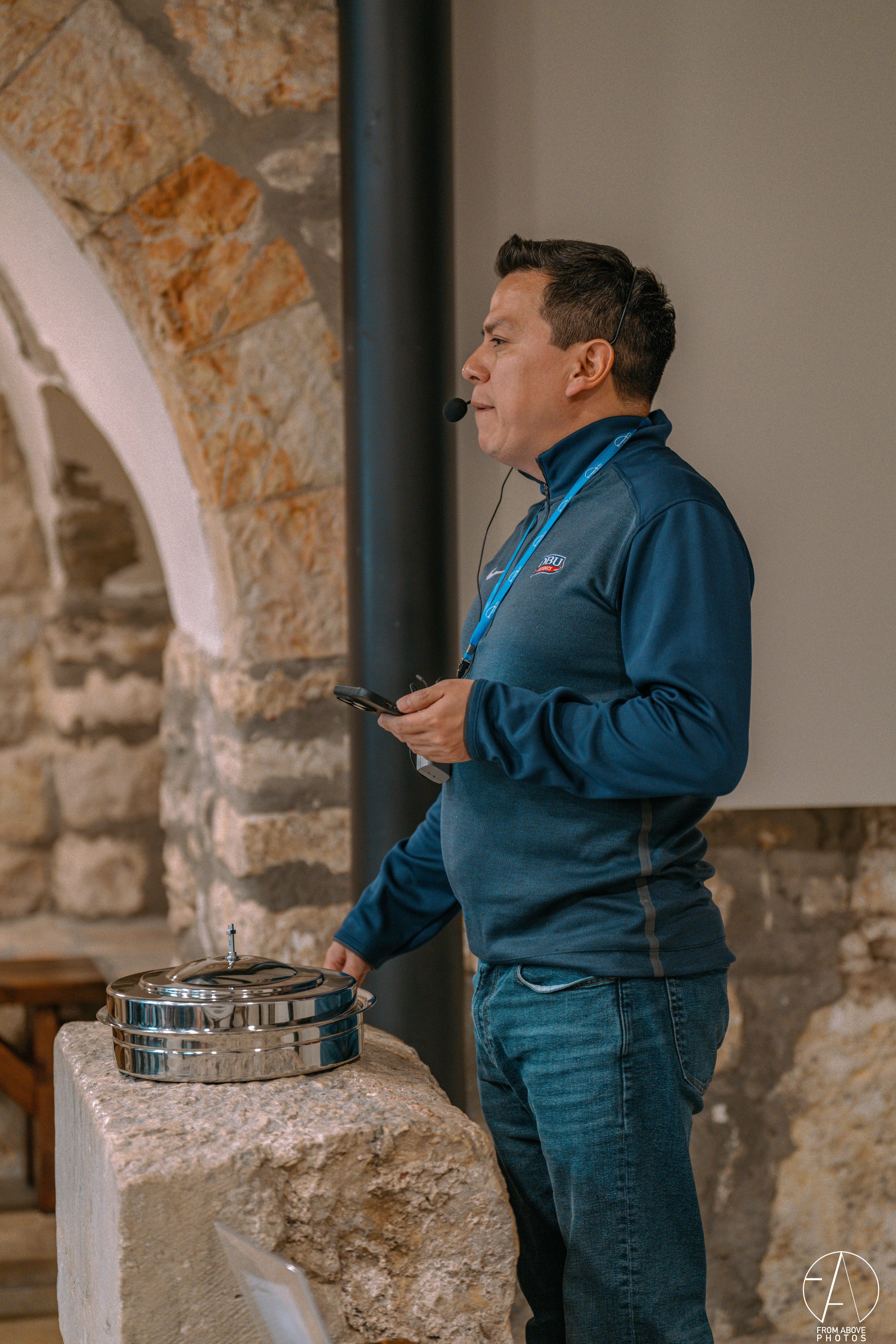 A man wearing a blue jacket with a logo, holding a phone, standing next to a stainless steel cake display, in a room with stone and brick walls.