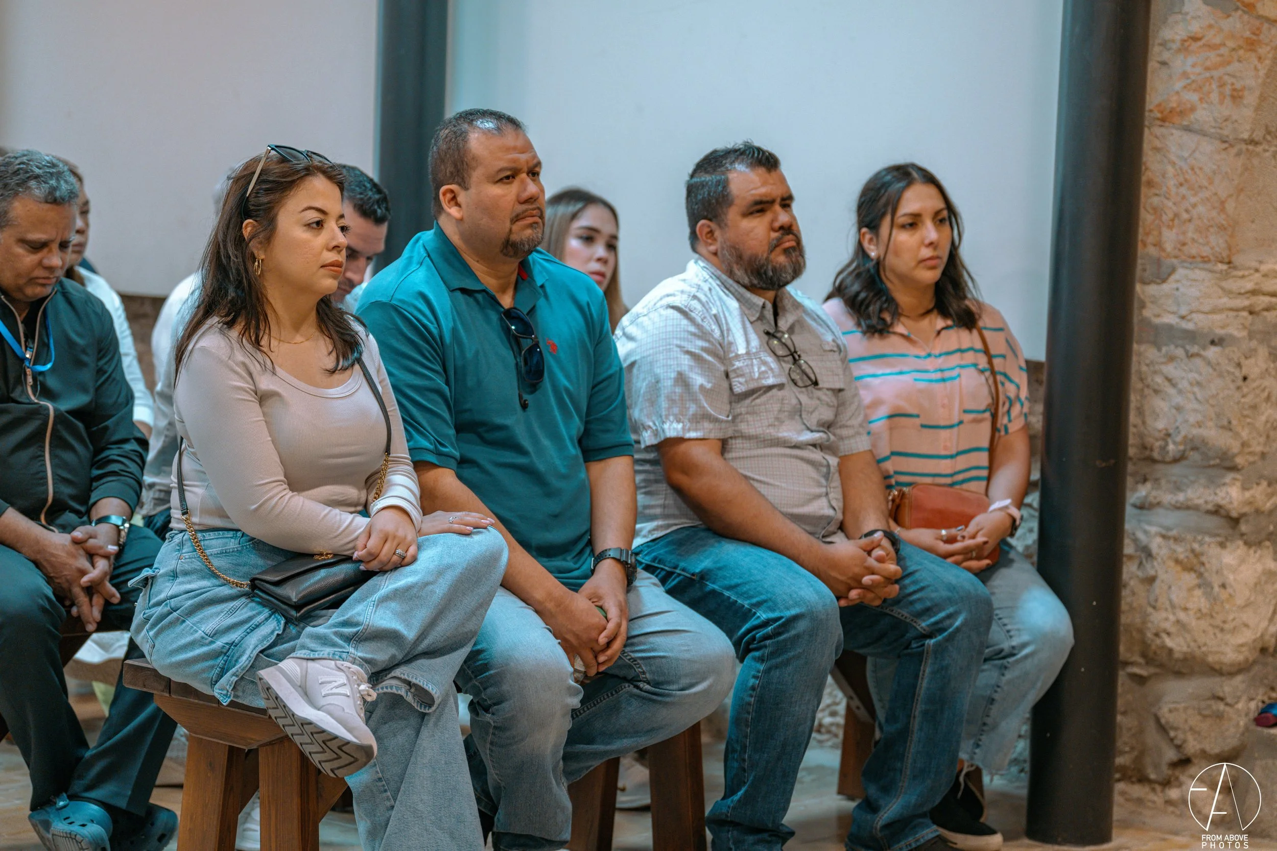 Group of people sitting and listening attentively at an indoor event or presentation, with a stone wall on the right.