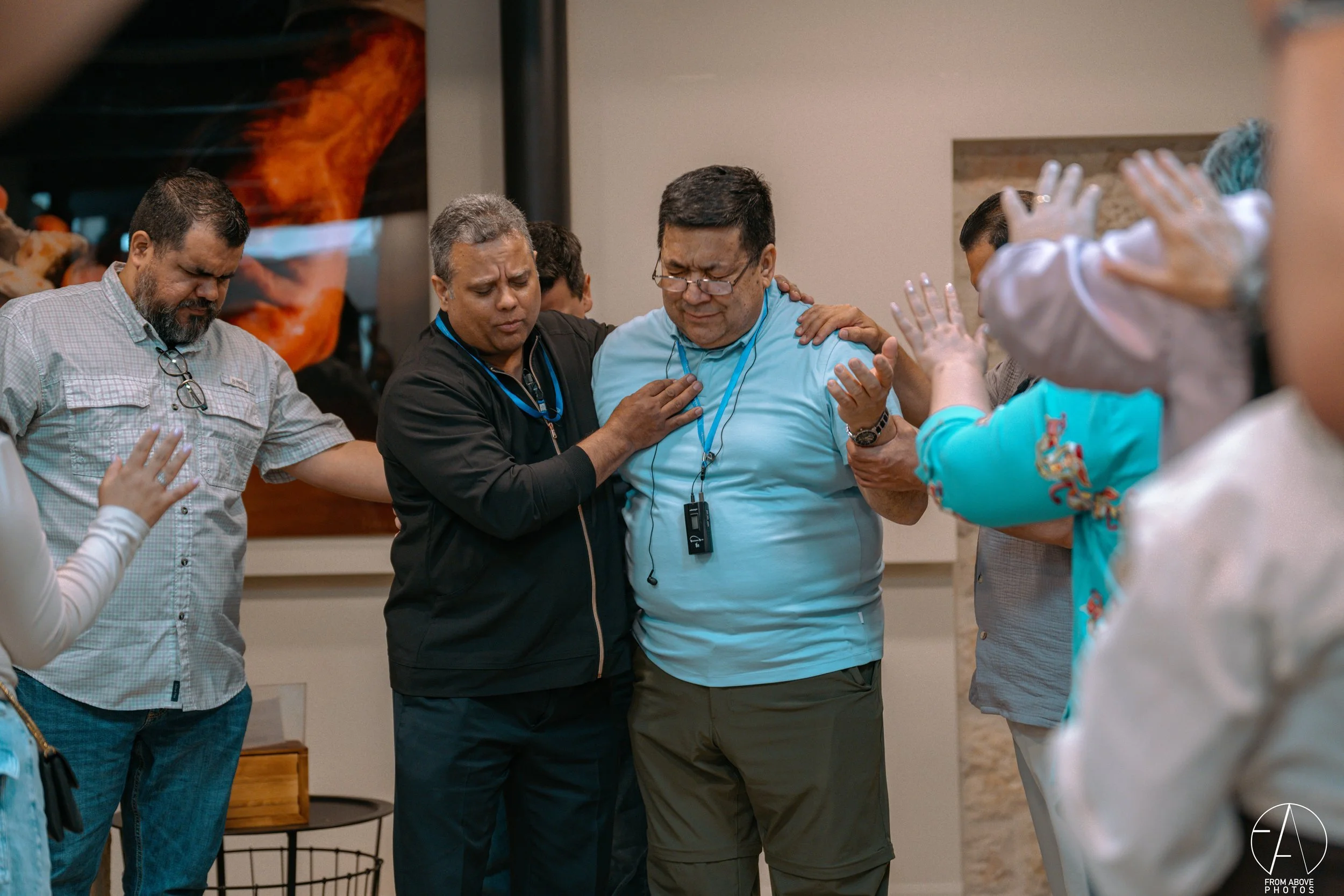 Group of people praying together with eyes closed and hands raised or placed on shoulders, some with eyes closed and hands raised.