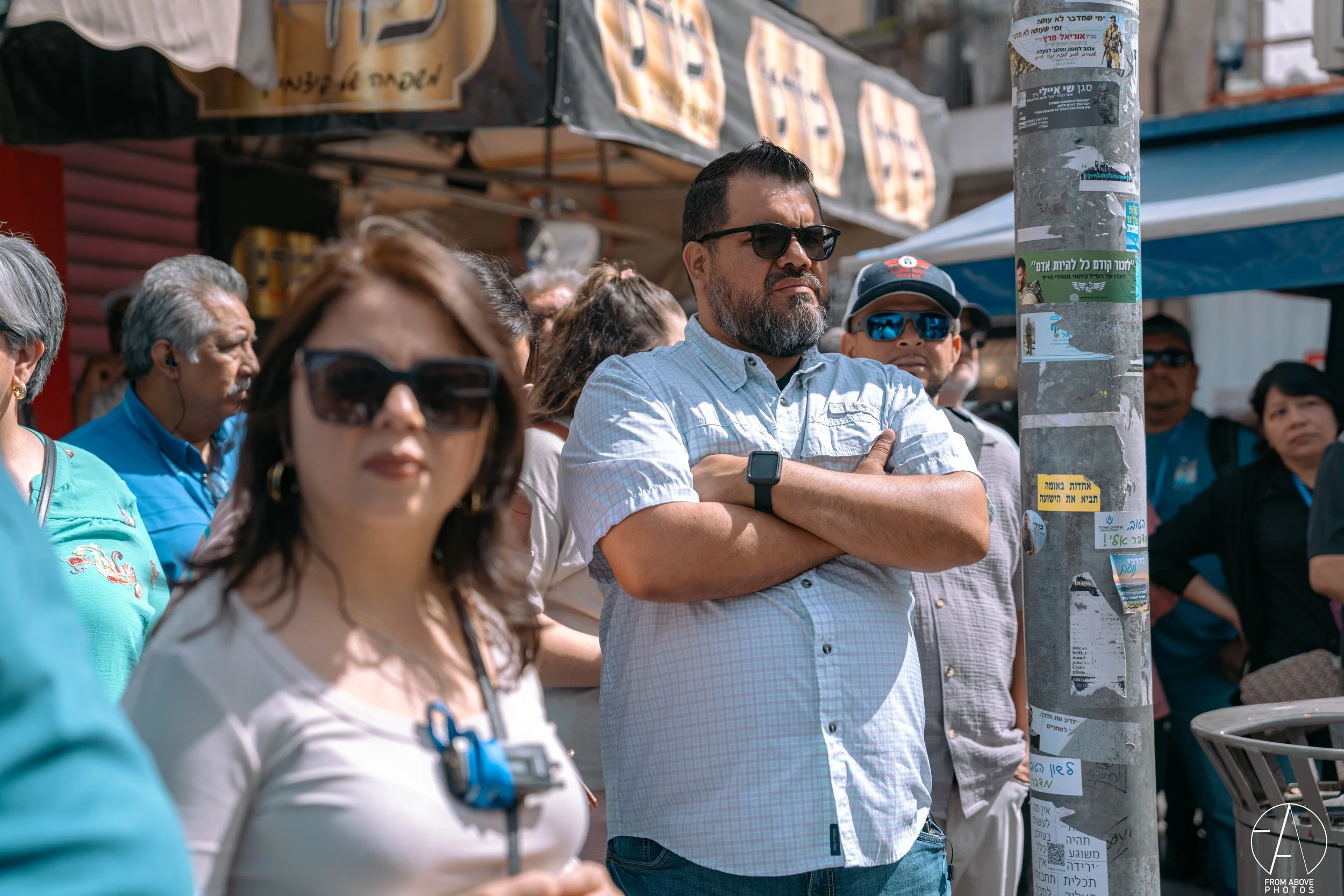 A group of diverse people standing outdoors in a crowd, some wearing sunglasses and casual clothing, listening attentively with a man in a blue checkered shirt crossing his arms in the center.