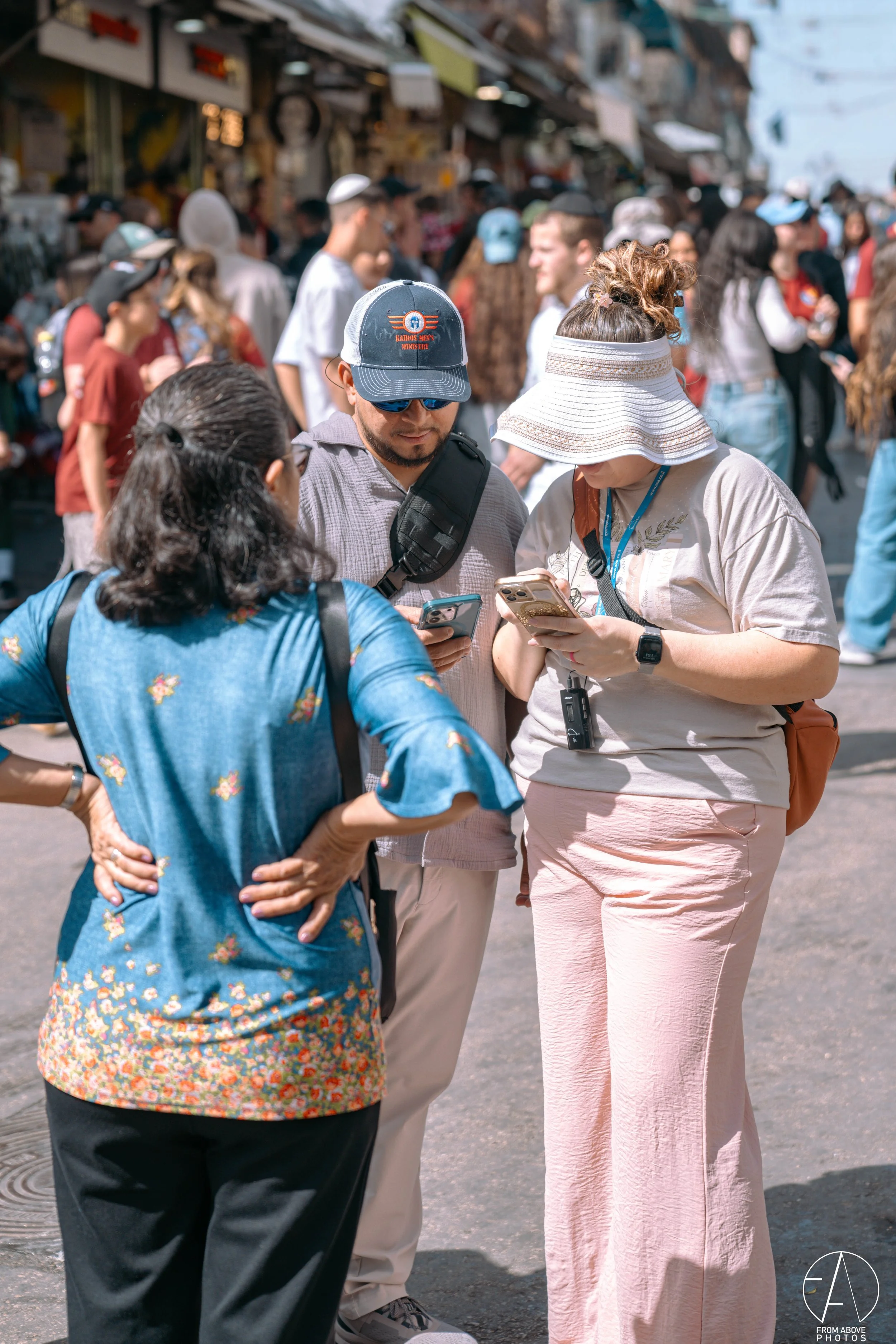Three people standing on a busy street, looking at their phones, with a crowd and market stalls in the background.