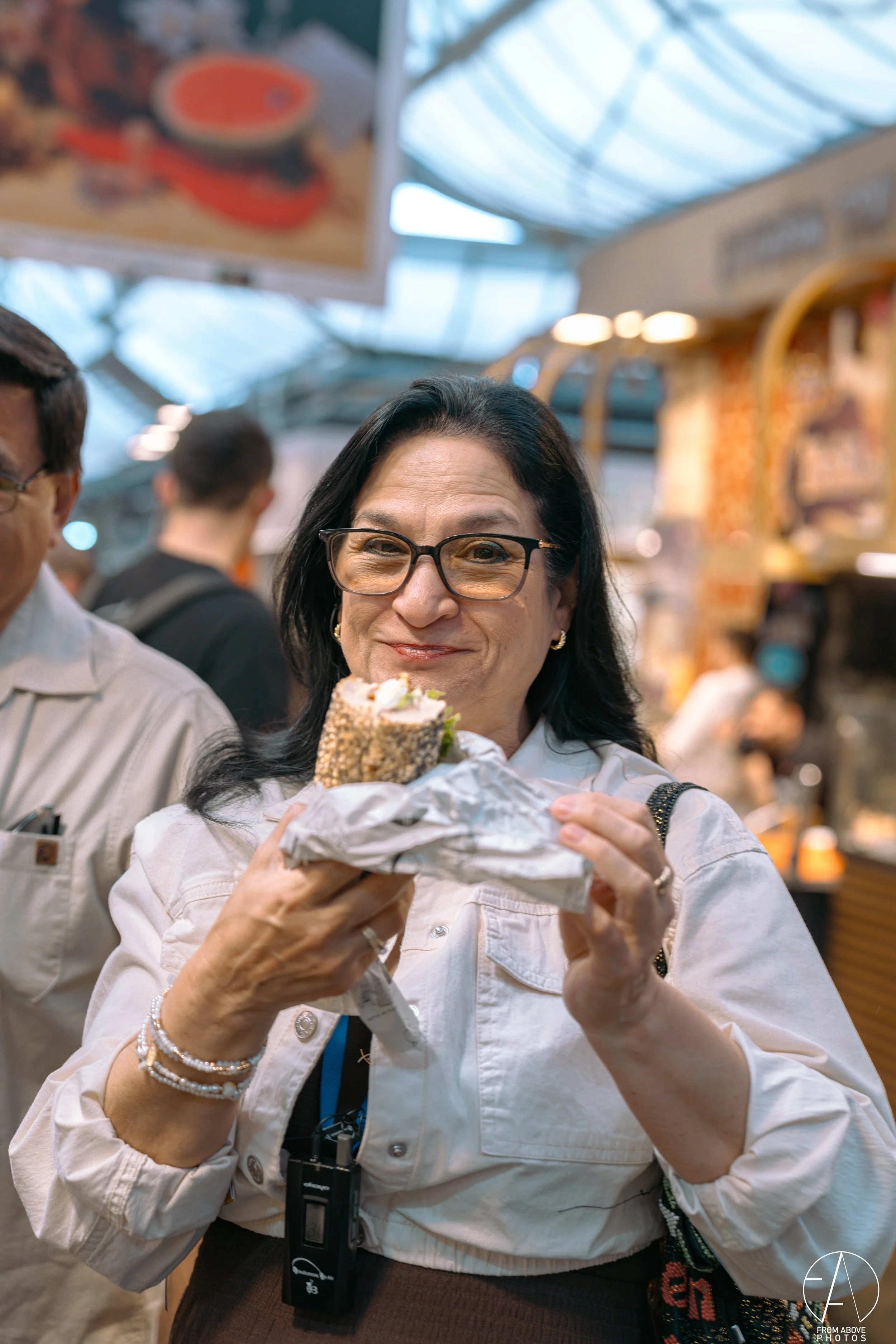 A smiling middle-aged woman with glasses holding a sandwich wrapped in foil in a busy indoor marketplace.