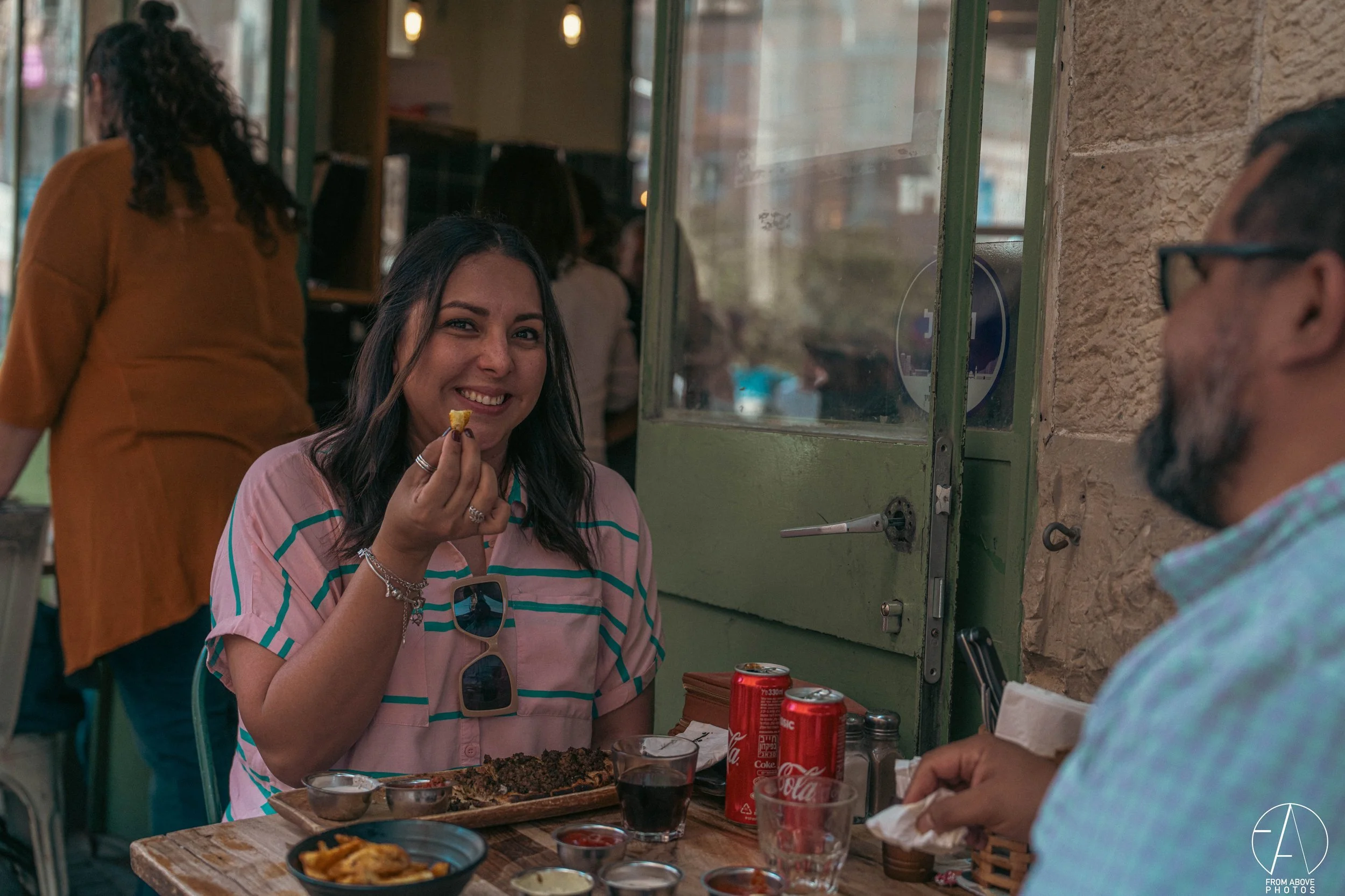 A woman with dark hair, wearing a pink and teal striped shirt and sunglasses hanging from her shirt, sits at a restaurant table holding a piece of food, smiling at the camera. There are two cans of Coca-Cola, a glass of soda, and various dishes on th