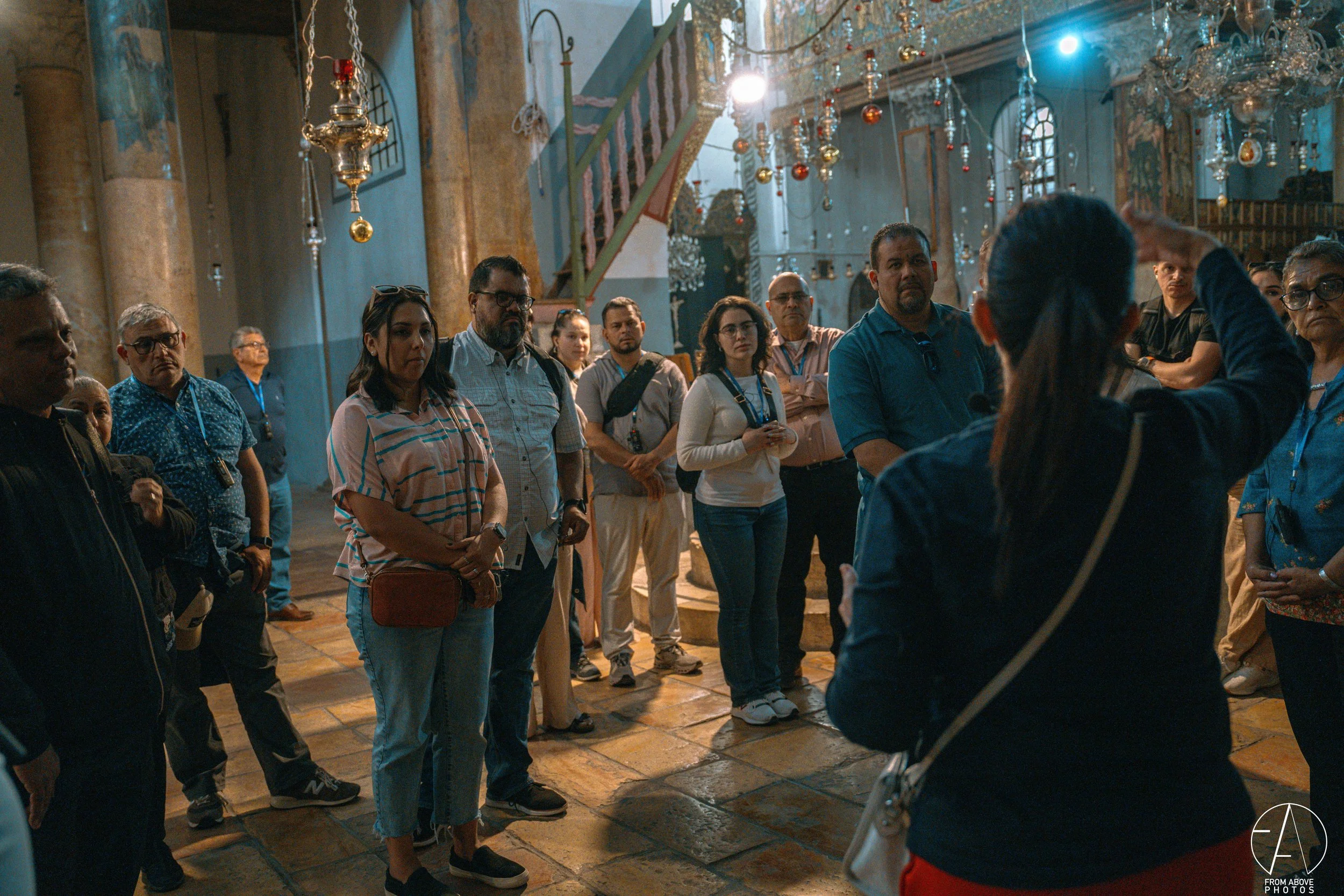 Tour group inside a church or cathedral, listening to a guide with her back to camera, surrounded by ornate chandeliers and religious decorations.